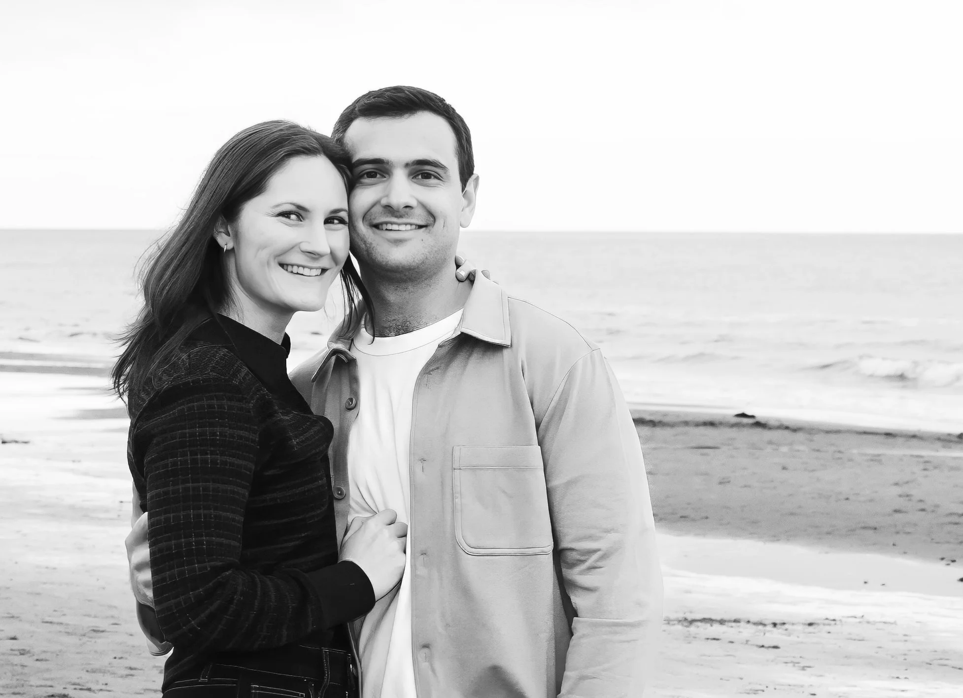 A smiling couple standing close together on a beach, with the ocean in the background. The woman has dark hair and wears a dark jacket, and the man has short dark hair and wears a light-colored jacket over a white shirt.