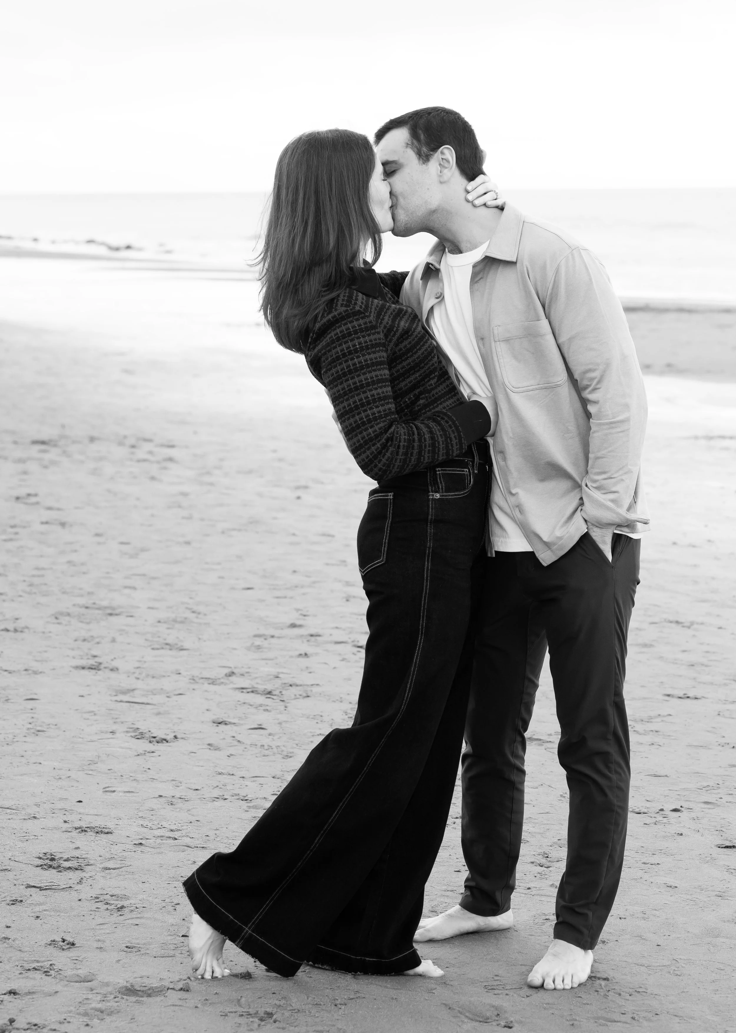 A black and white photo of a couple kissing at the beach, with the woman in a long skirt and she is barefoot, and the man standing with his hand behind her back, both barefoot on the sand.
