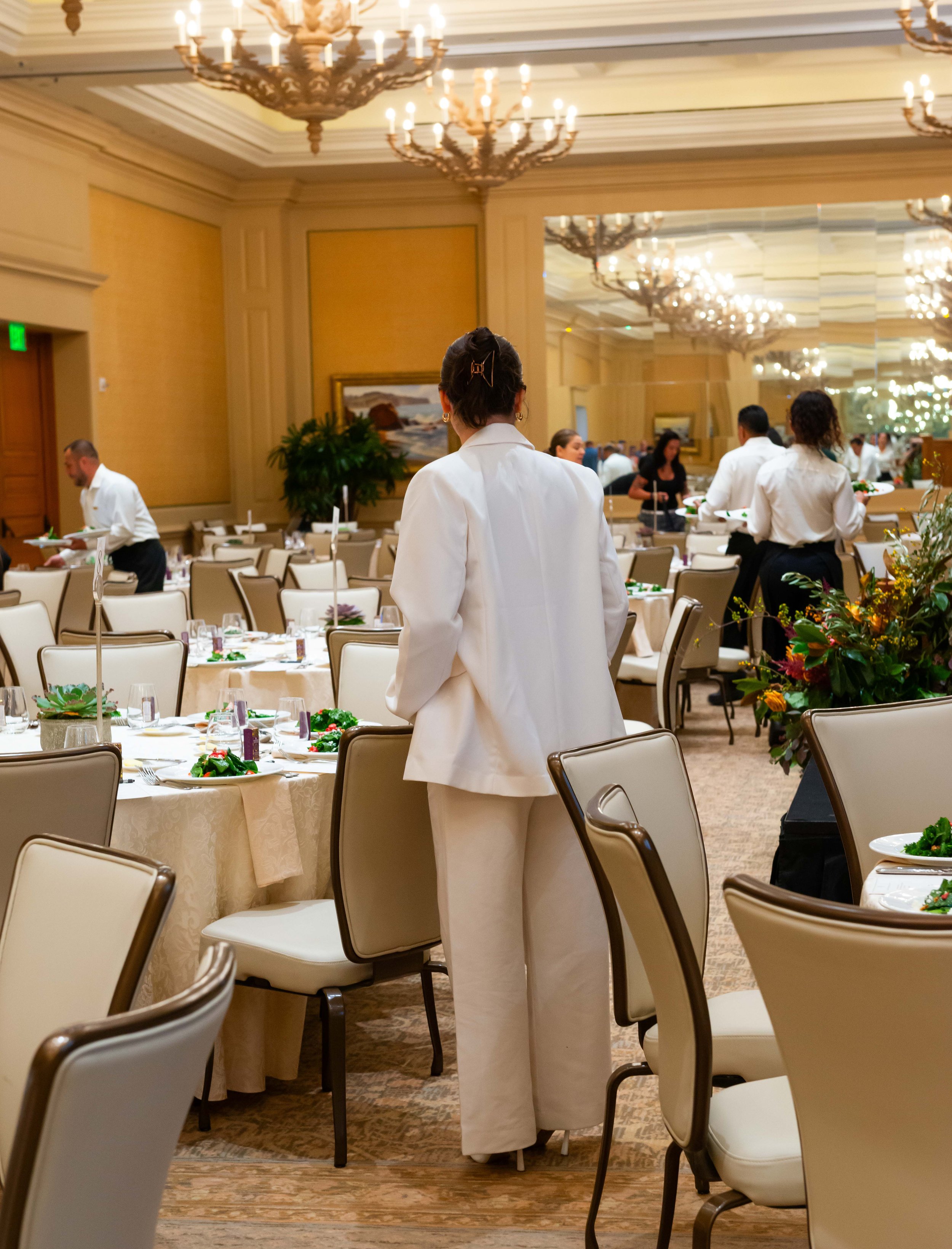Waitstaff setting tables in an elegant banquet hall with chandeliers and floral centerpieces.