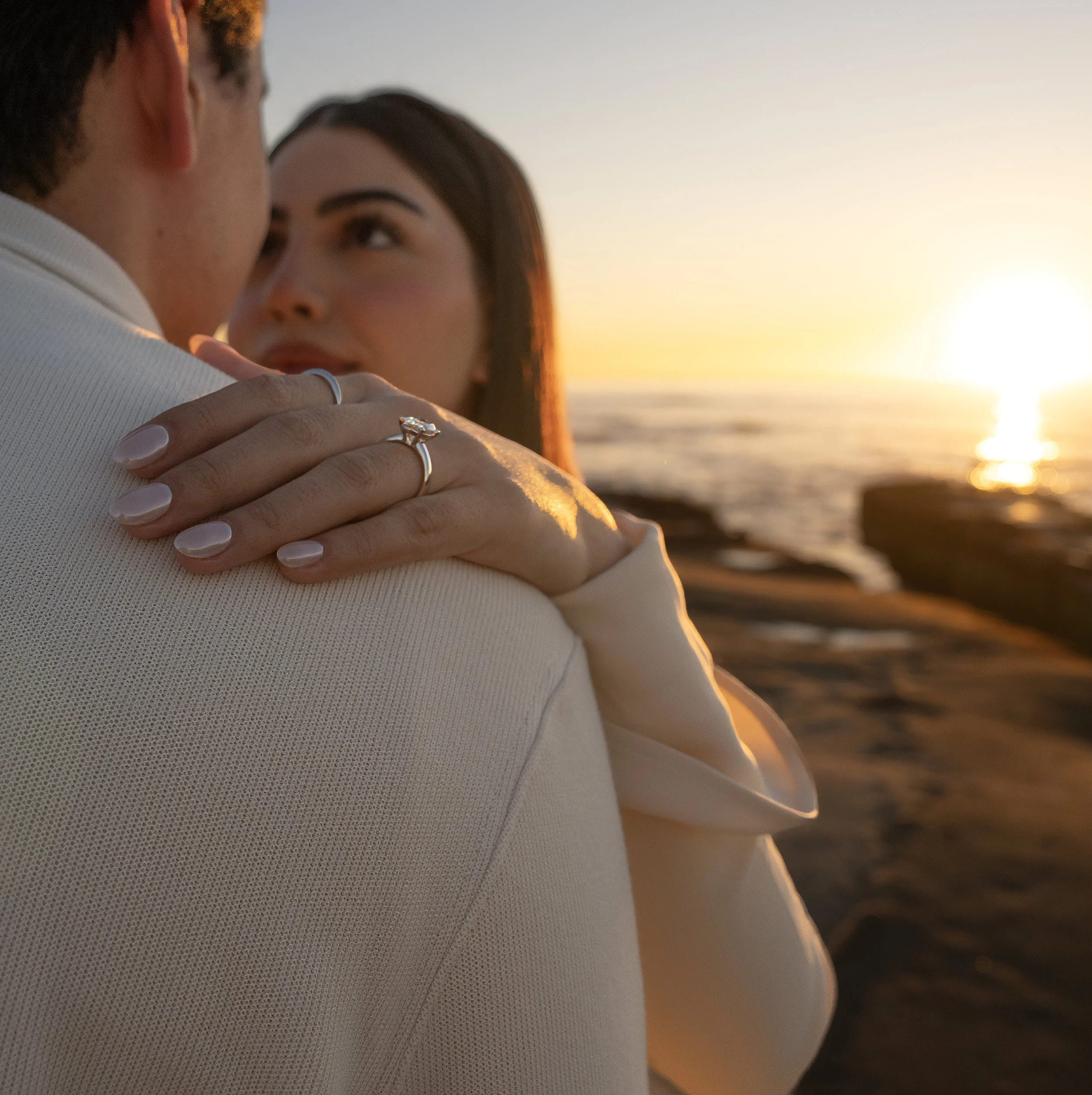 A couple hugging during sunset, with the woman's face visible and the man’s face partially obscured. The woman has dark hair and is wearing a ring on her finger. The background features a setting sun over water, creating a warm glow.