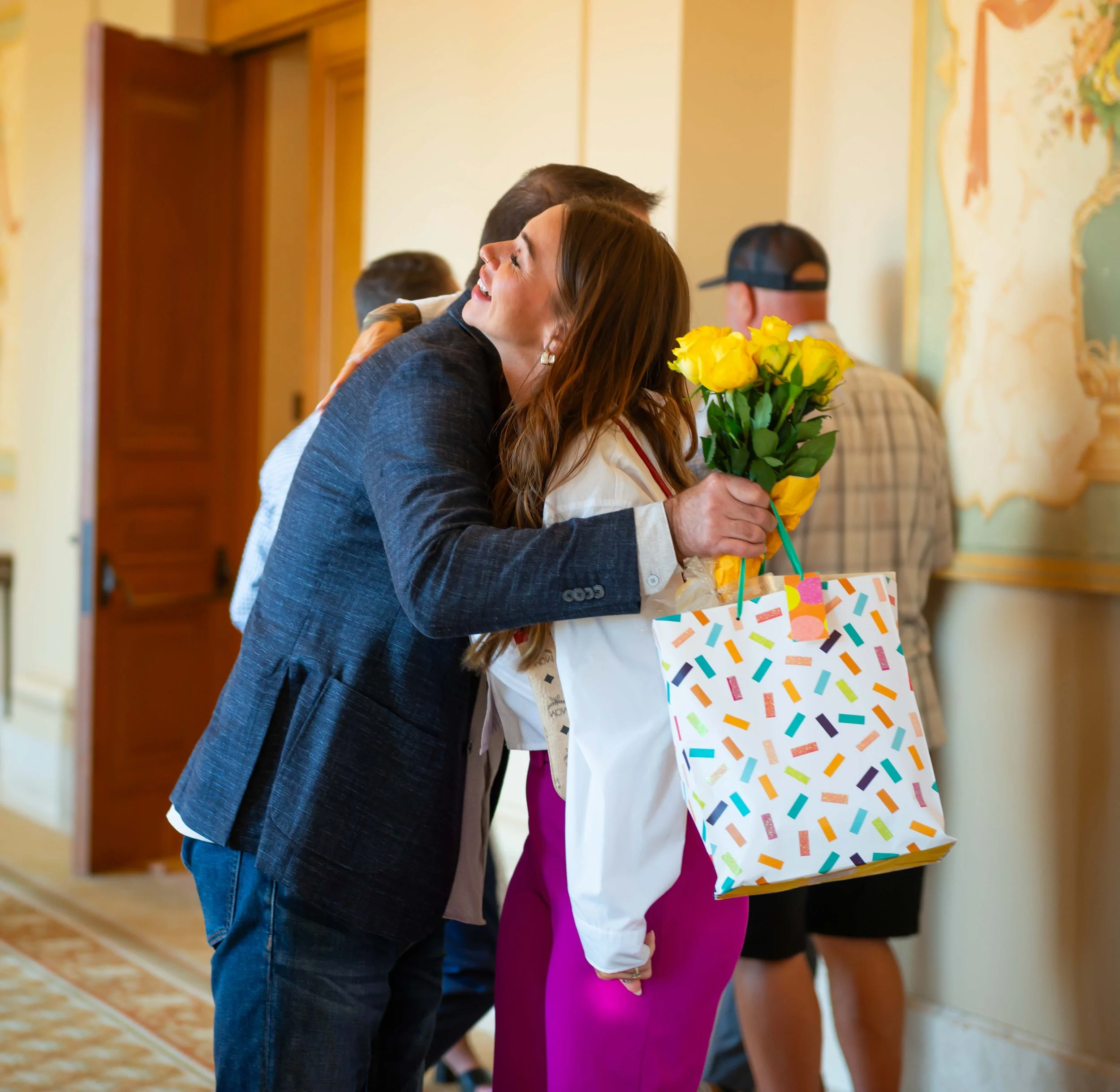 People standing in line, hugging, and holding a bouquet of yellow roses at an indoor event.