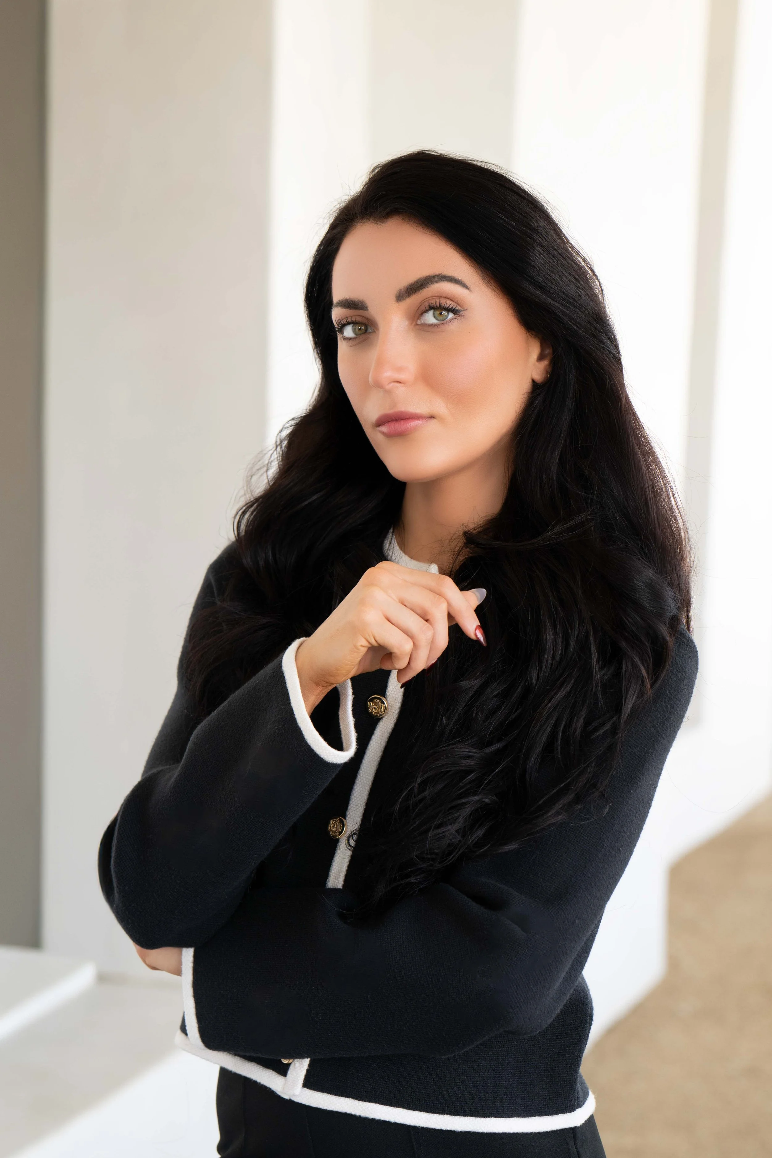 Portrait of a woman with dark hair and light green eyes standing indoors near a wall with indirect sunlight, wearing a black and white cardigan and a white shirt, with her hand near her chin.