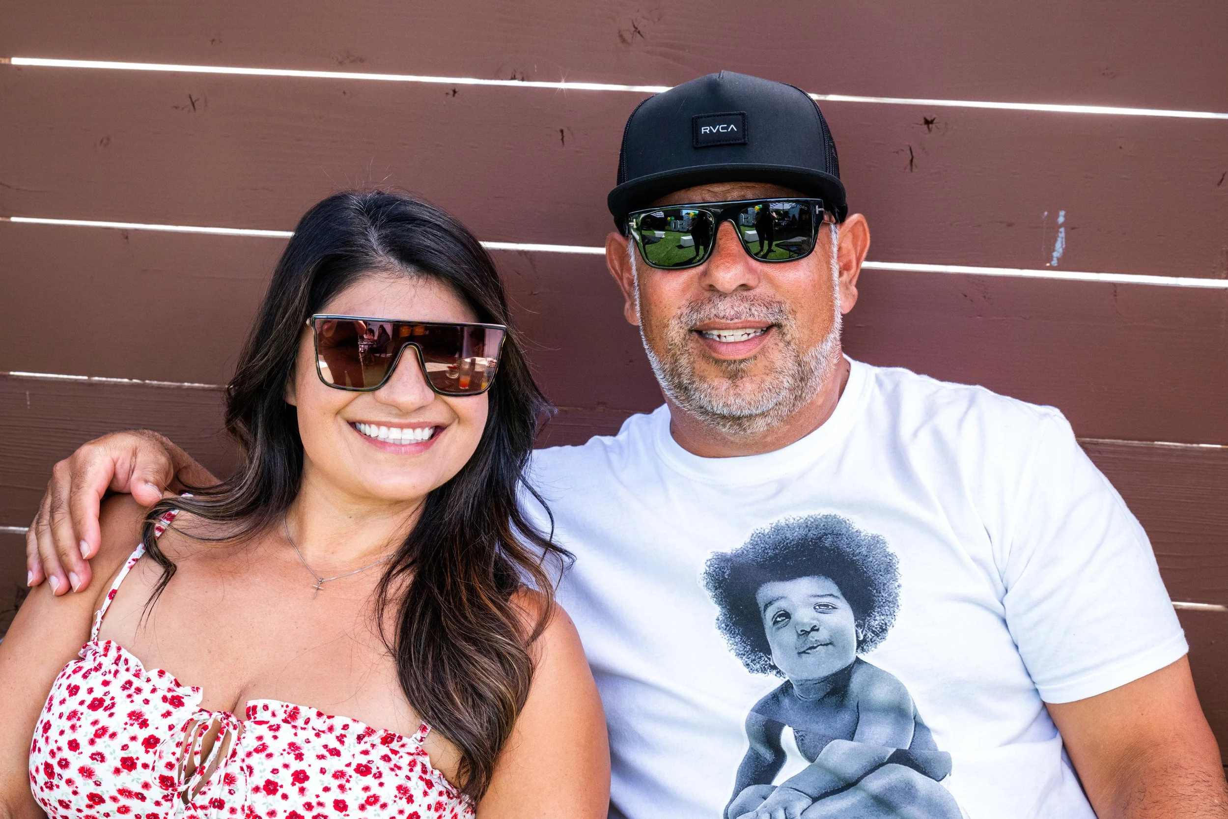 A smiling woman and a smiling man taking a selfie against a wooden fence, both wearing sunglasses.