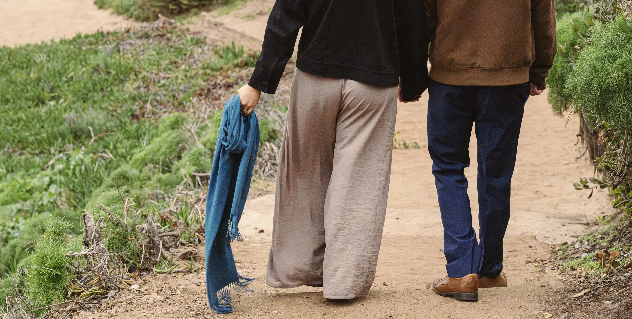Two people walking hand in hand on a dirt path, one carrying a blue scarf.