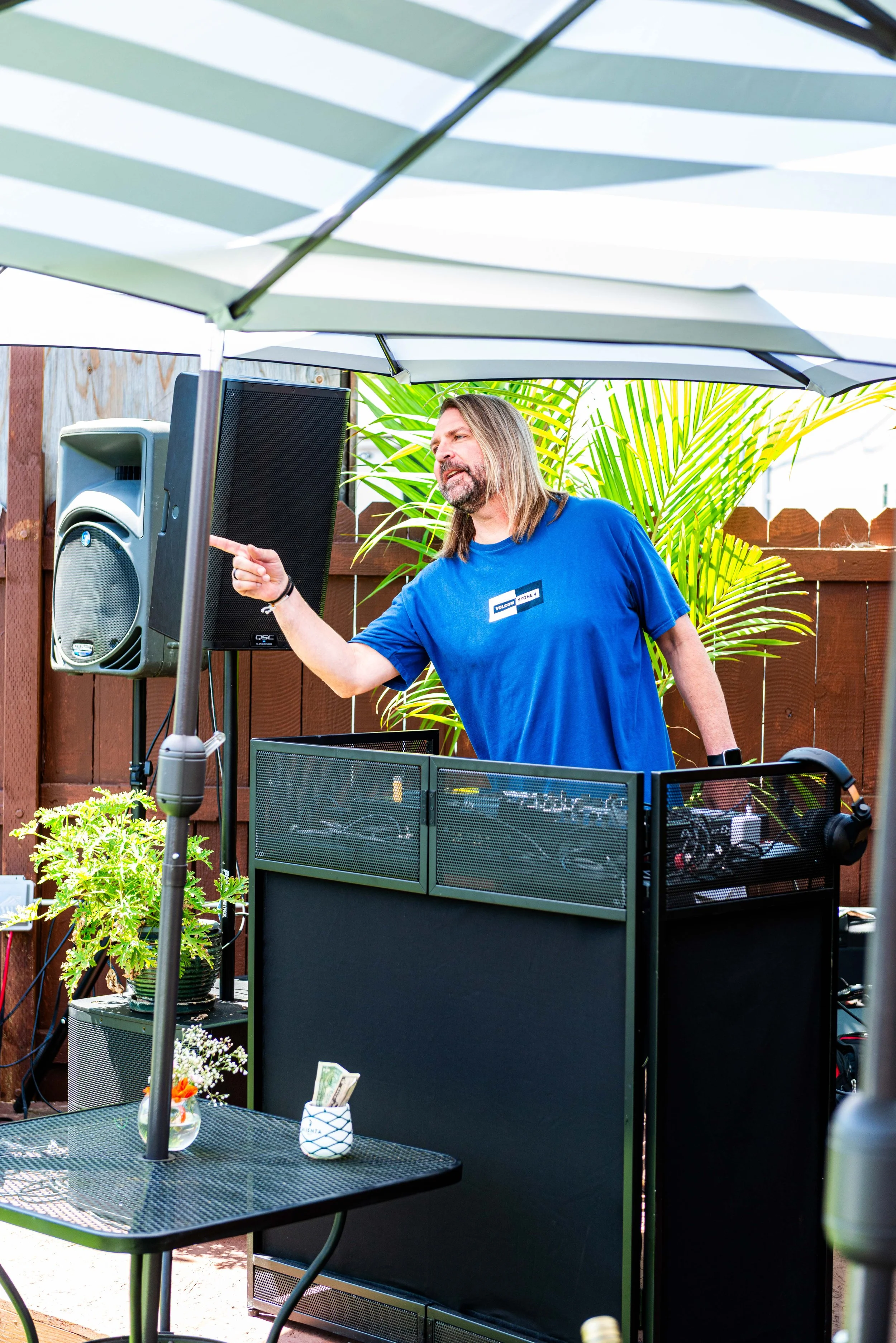 A man with long gray hair and a beard, wearing a blue T-shirt, pointing at a large speaker, standing outdoors beneath a patio umbrella, with a wooden fence, green plants, and a table with flowers and a cup in the background.