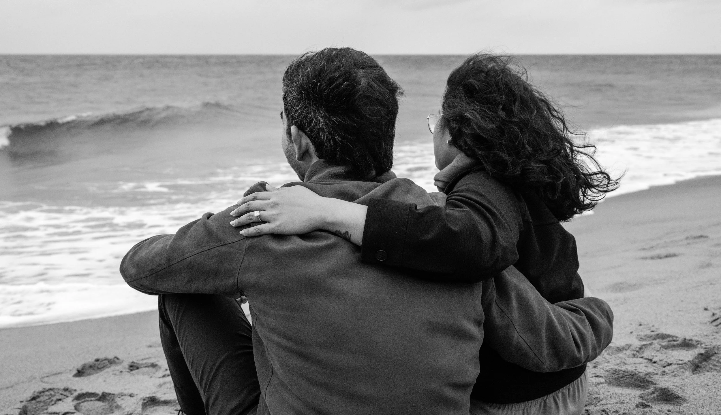 Two people sitting on a beach embracing each other, with waves in the background, black and white photograph.