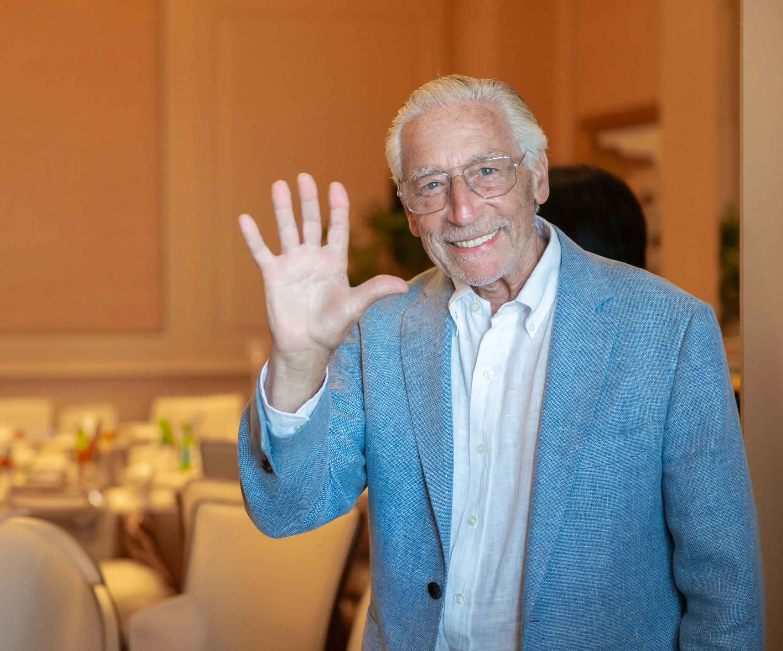 An elderly man with gray hair, wearing glasses, a white shirt, and a light blue blazer, smiling and waving.