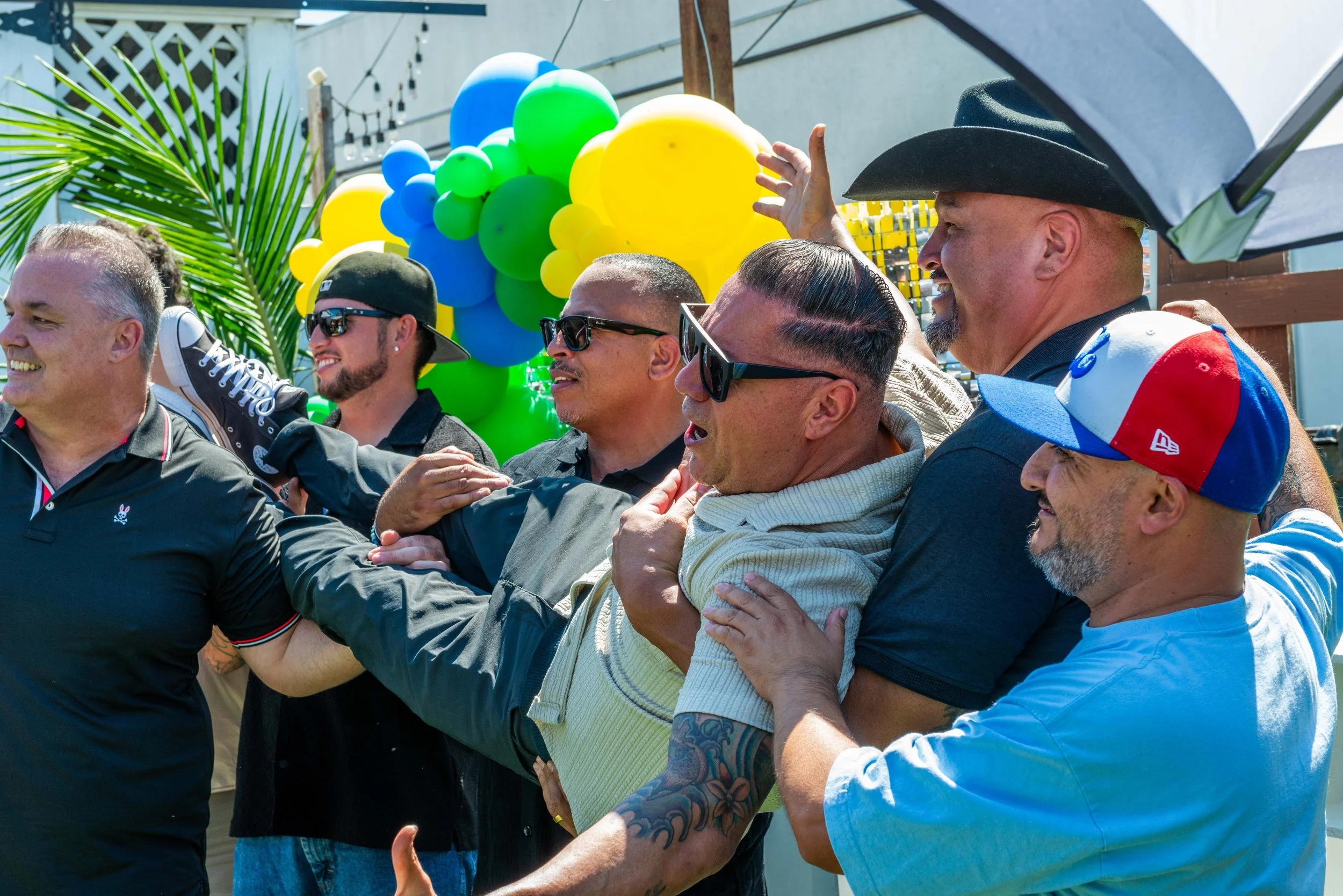 A group of people participating in a celebratory event, forming a human chain with their hands on each other's shoulders, with colorful balloons and palm leaves in the background.