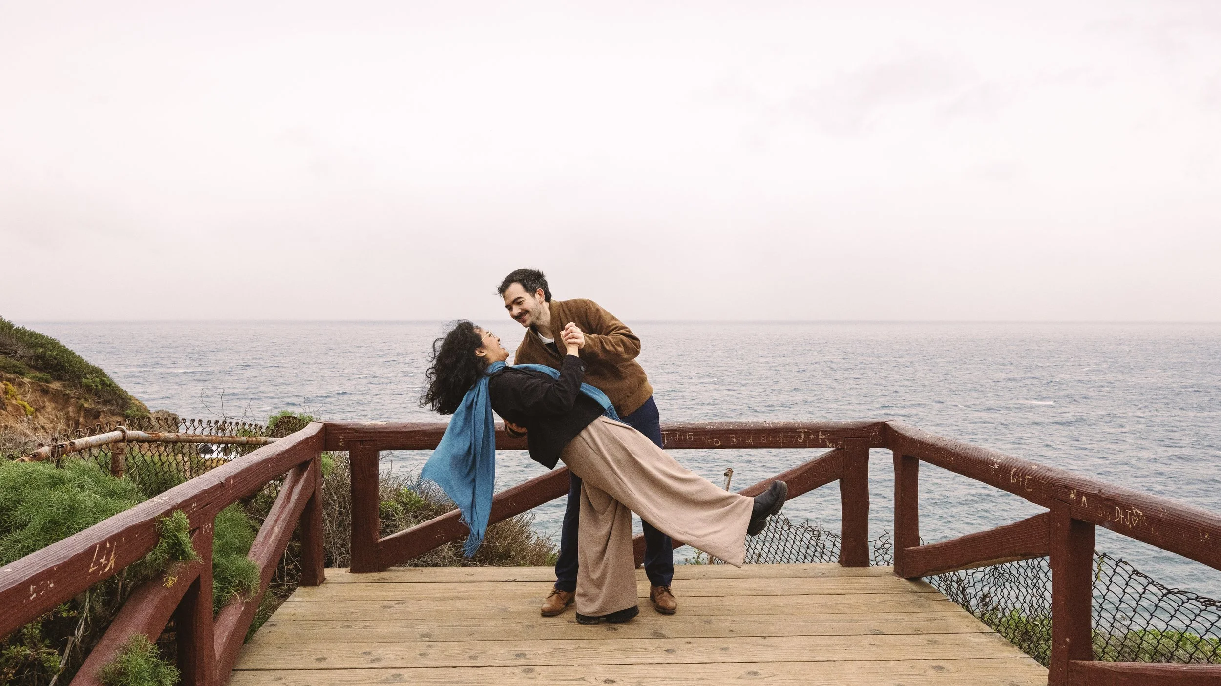 A couple dancing on a wooden platform by the seaside with overcast sky, holding hands and smiling at each other.