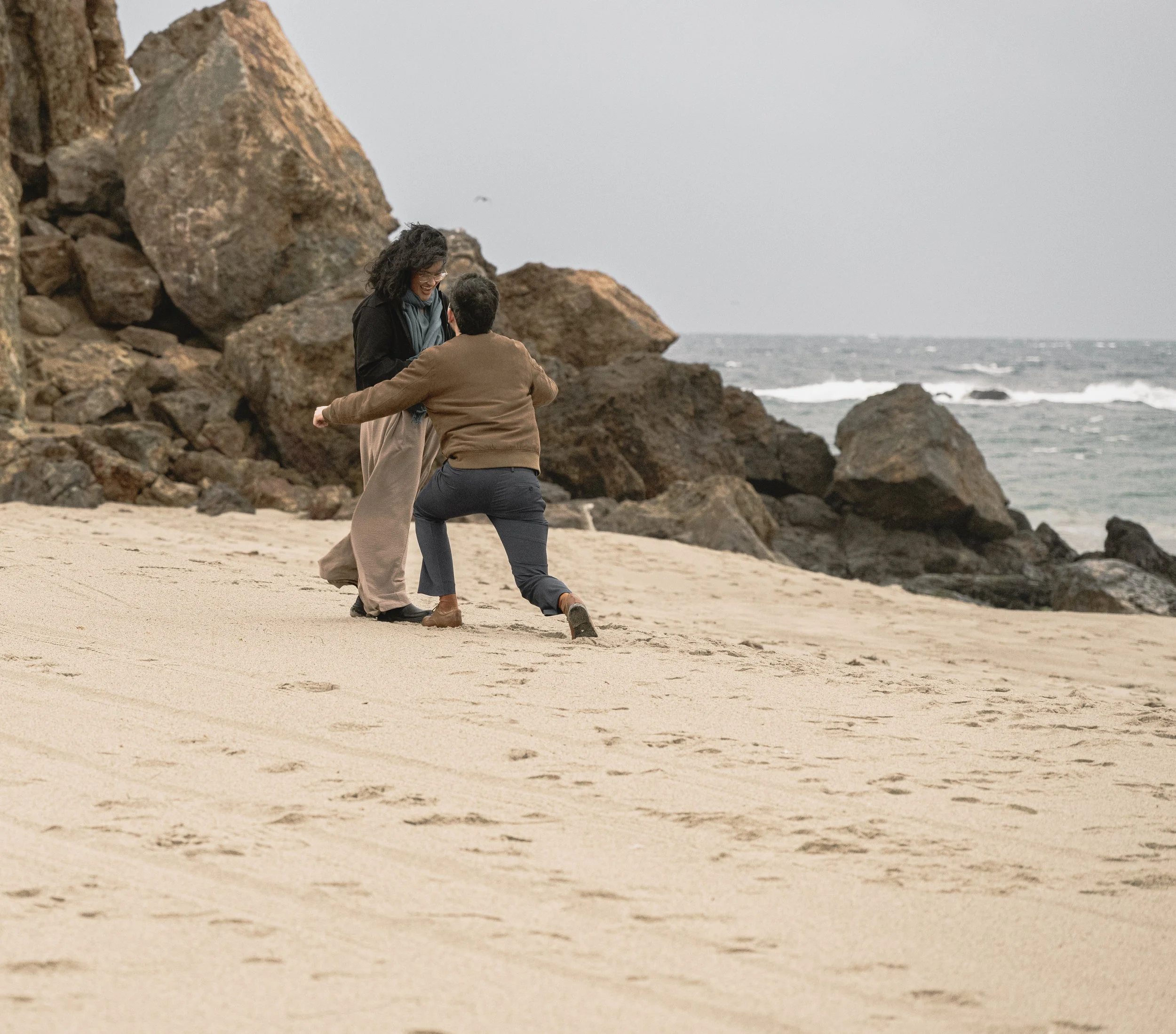 A man and a woman smiling and holding hands on a sandy beach with large rocks and the ocean in the background.