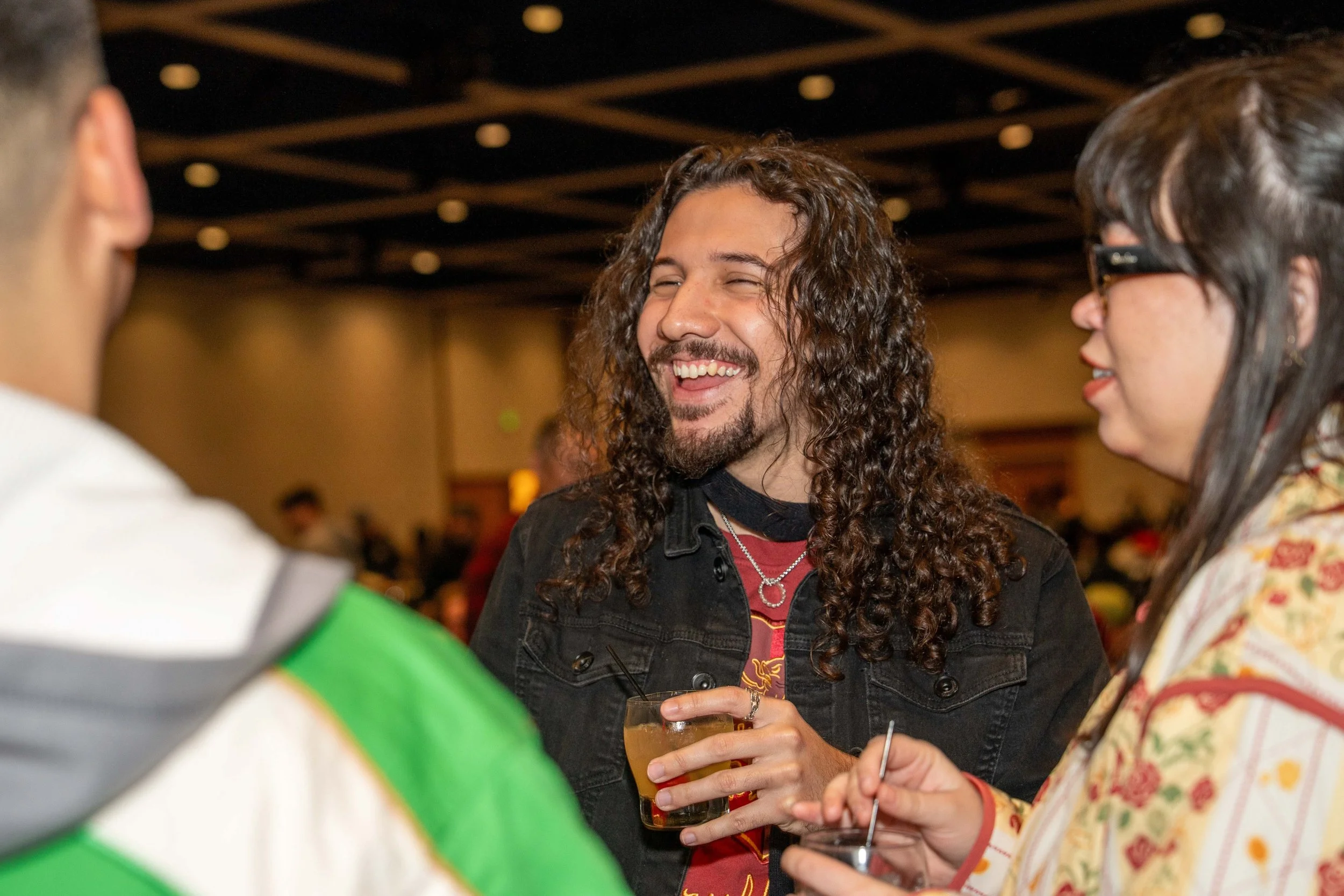 A group of three people socializing at an indoor event, including a man with long curly hair and a beard holding a drink, engaged in conversation with a woman wearing glasses and a patterned blazer, while another person is partially visible in the fo