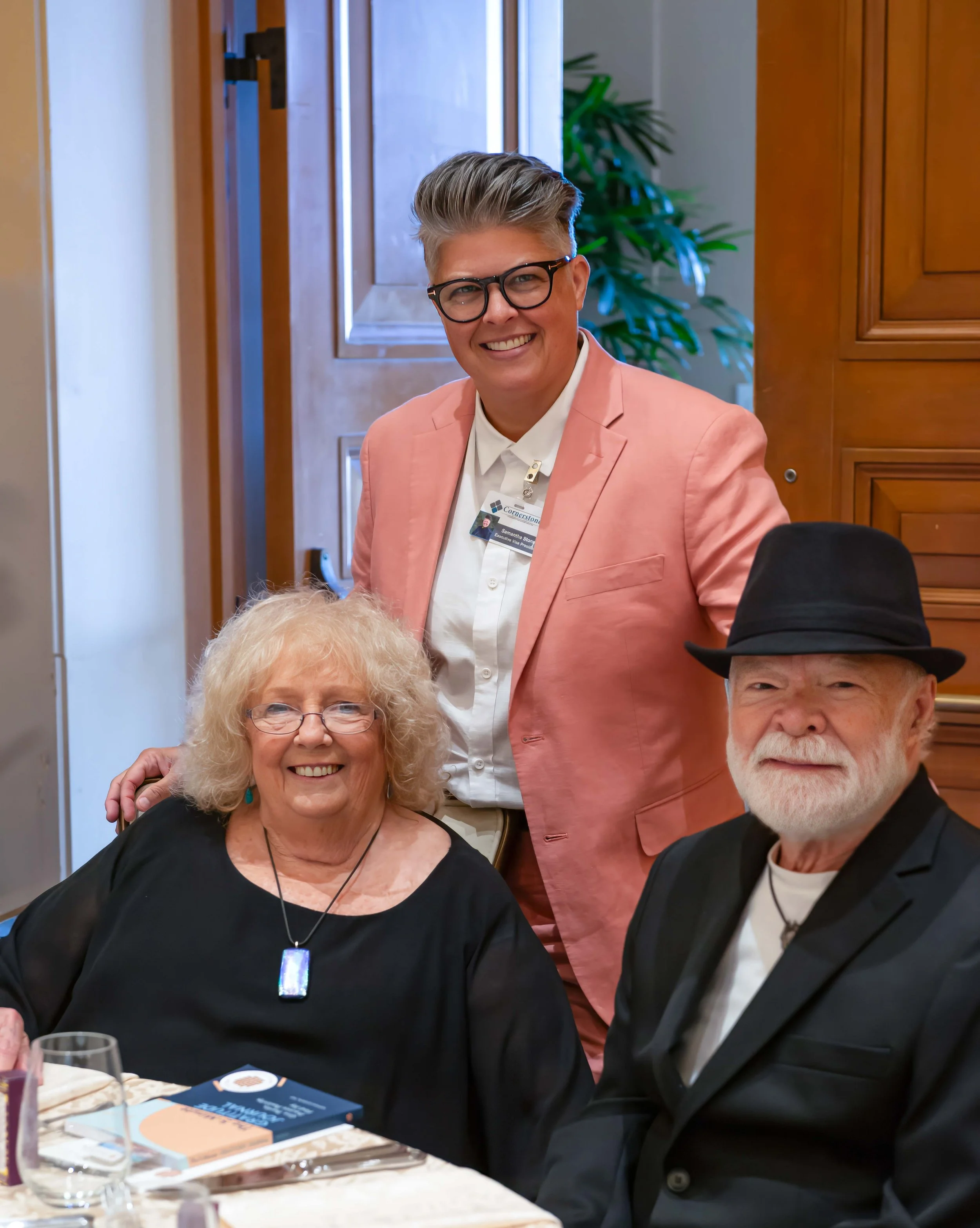 A smiling woman with glasses and curly blonde hair sitting at a table with a woman speaking with gray hair and a man with glasses and a beard. The woman is wearing a black top and a necklace with a pendant, and there is a book titled 'The Confidence'