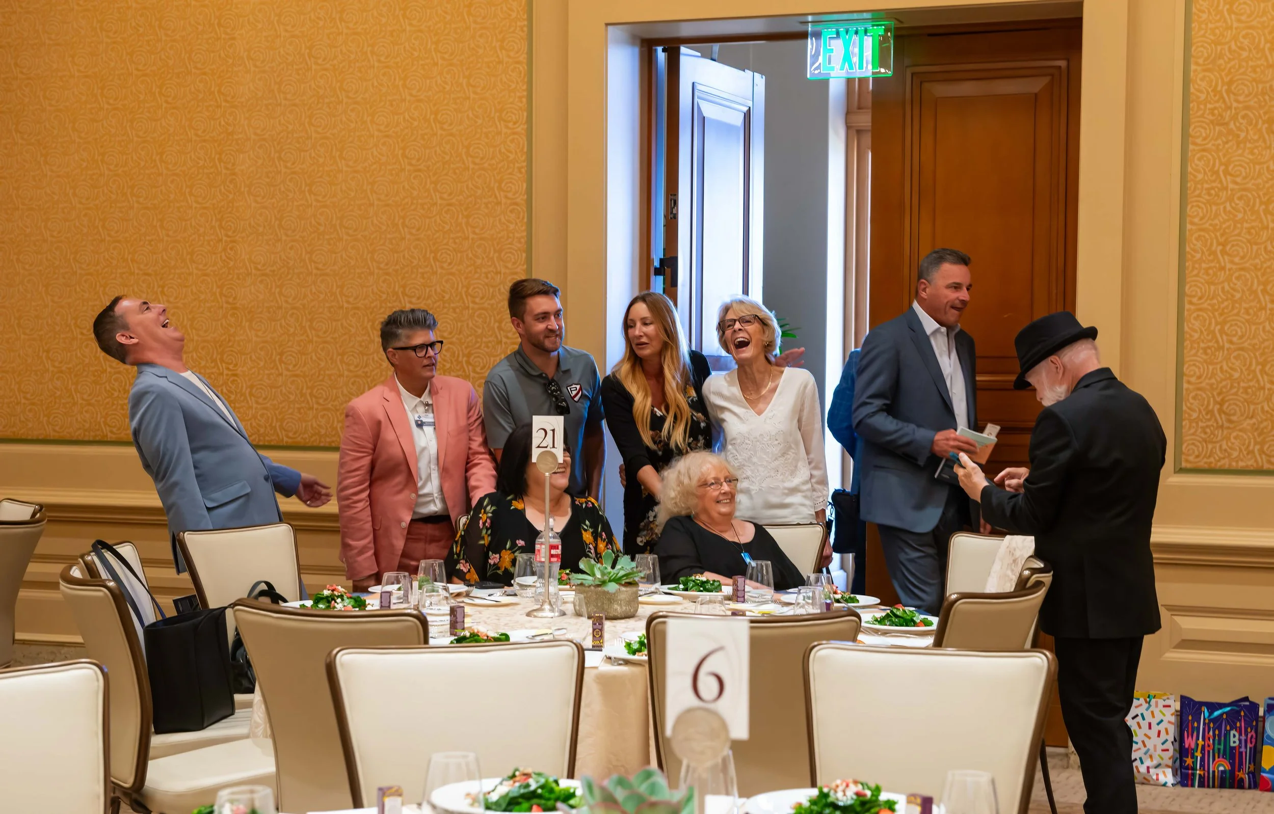 Group of people celebrating at a banquet, some standing and some seated at a decorated table, with an exit sign above and a warm yellow wall in the background.