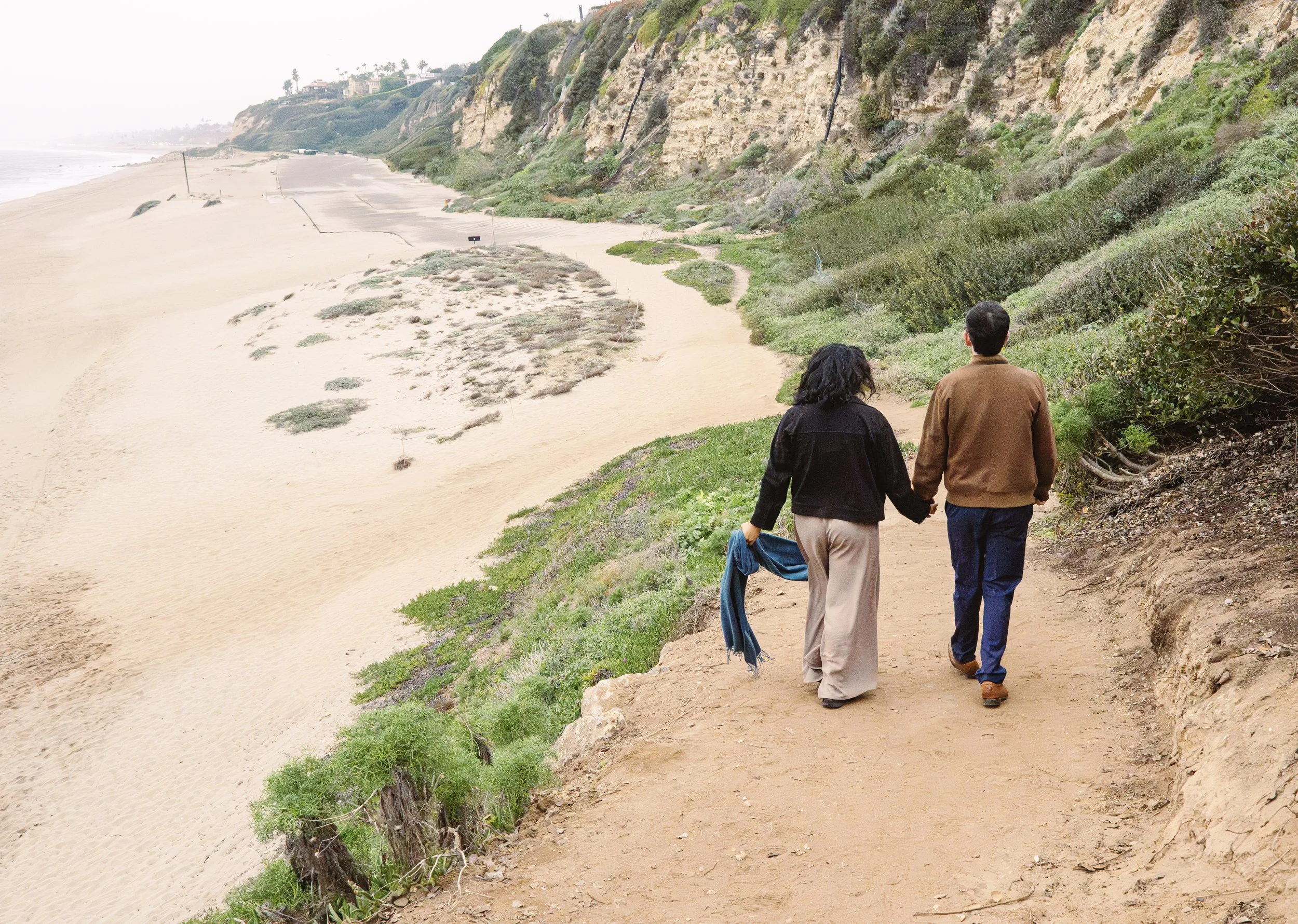Two people, a woman and a man, walking along a coastal trail with sandy beach and green vegetation on one side and tall cliffs on the other.