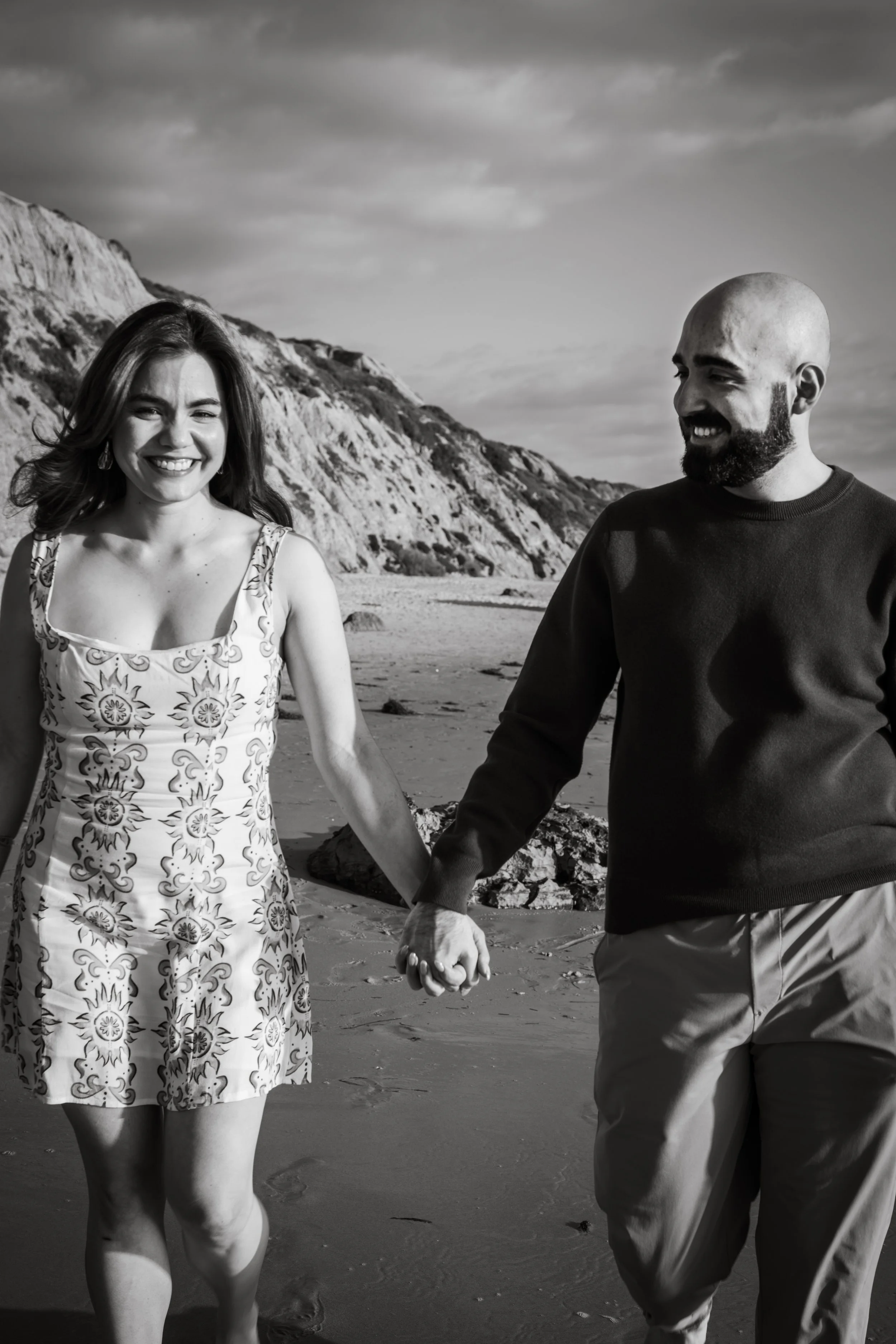 A smiling woman and man holding hands and walking on a beach with cliffs in the background, in black and white.