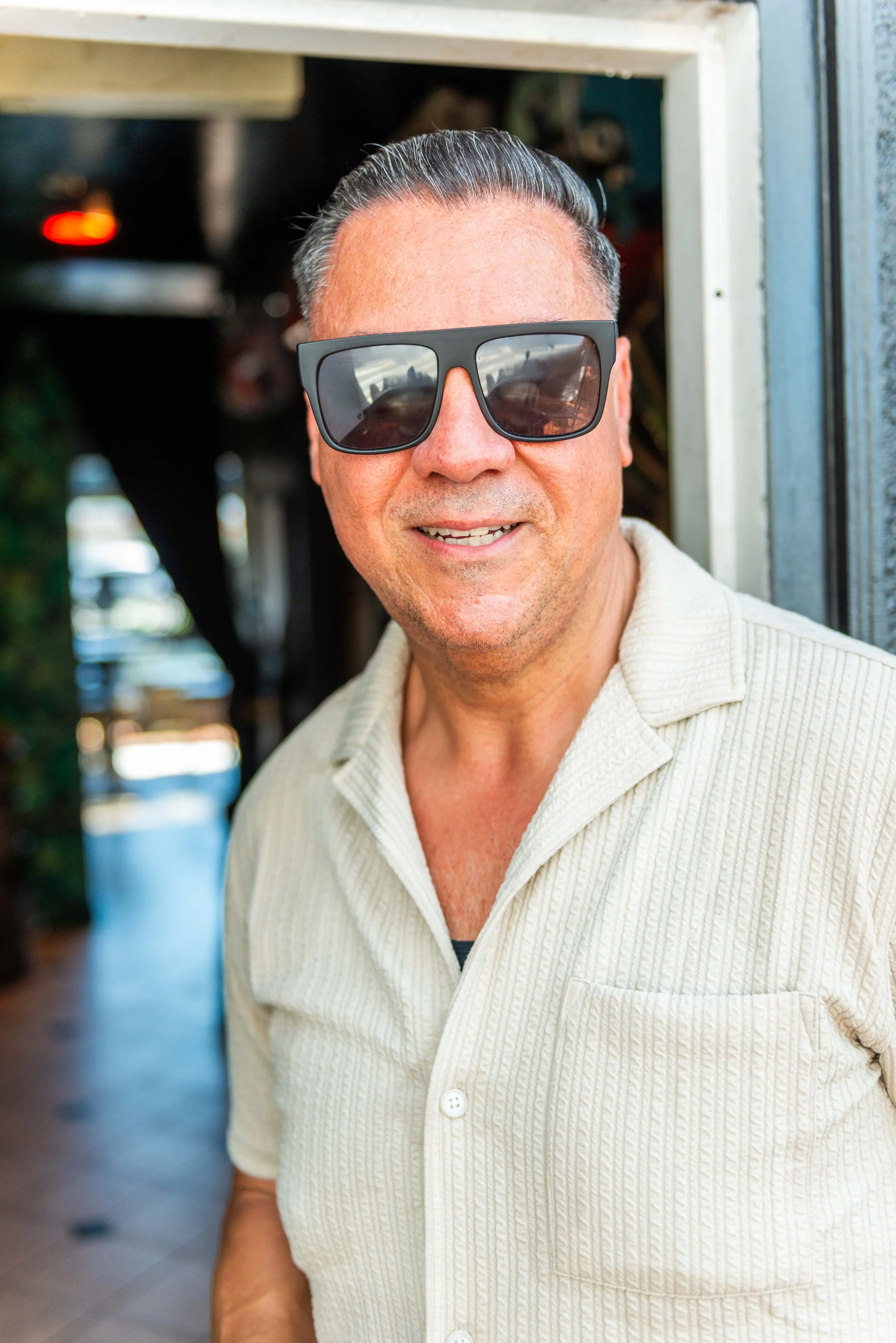 A middle-aged man with short, slicked-back dark hair, wearing large black sunglasses and a cream-colored, textured short-sleeve button-up shirt, smiling indoors with blurred background.