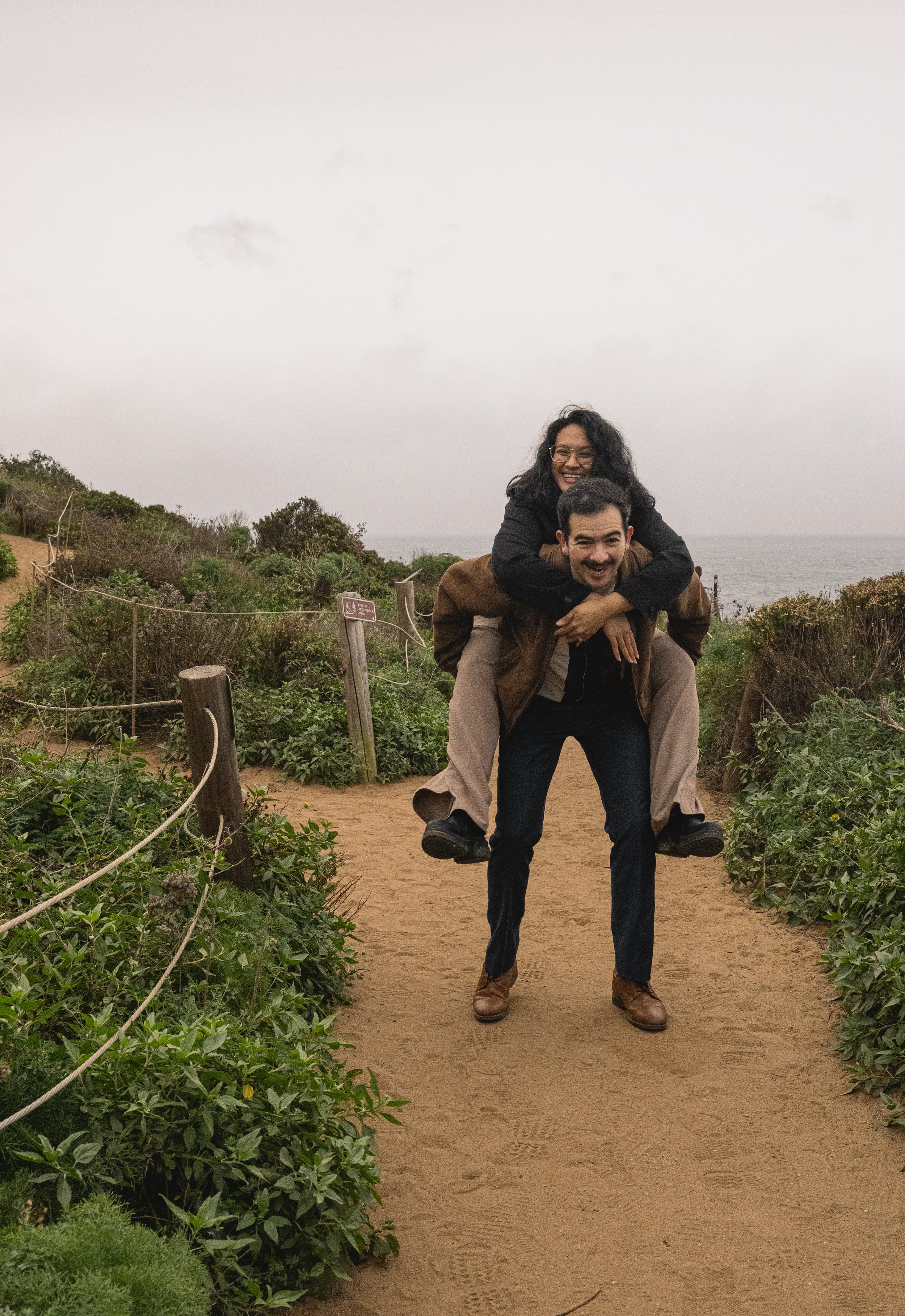 Two people, a woman and a man, playing on a sandy path outdoors near the coast. The woman is riding on the man's back, and both are smiling and enjoying themselves. The scene is set on a cloudy day, with greenery and the ocean in the background.