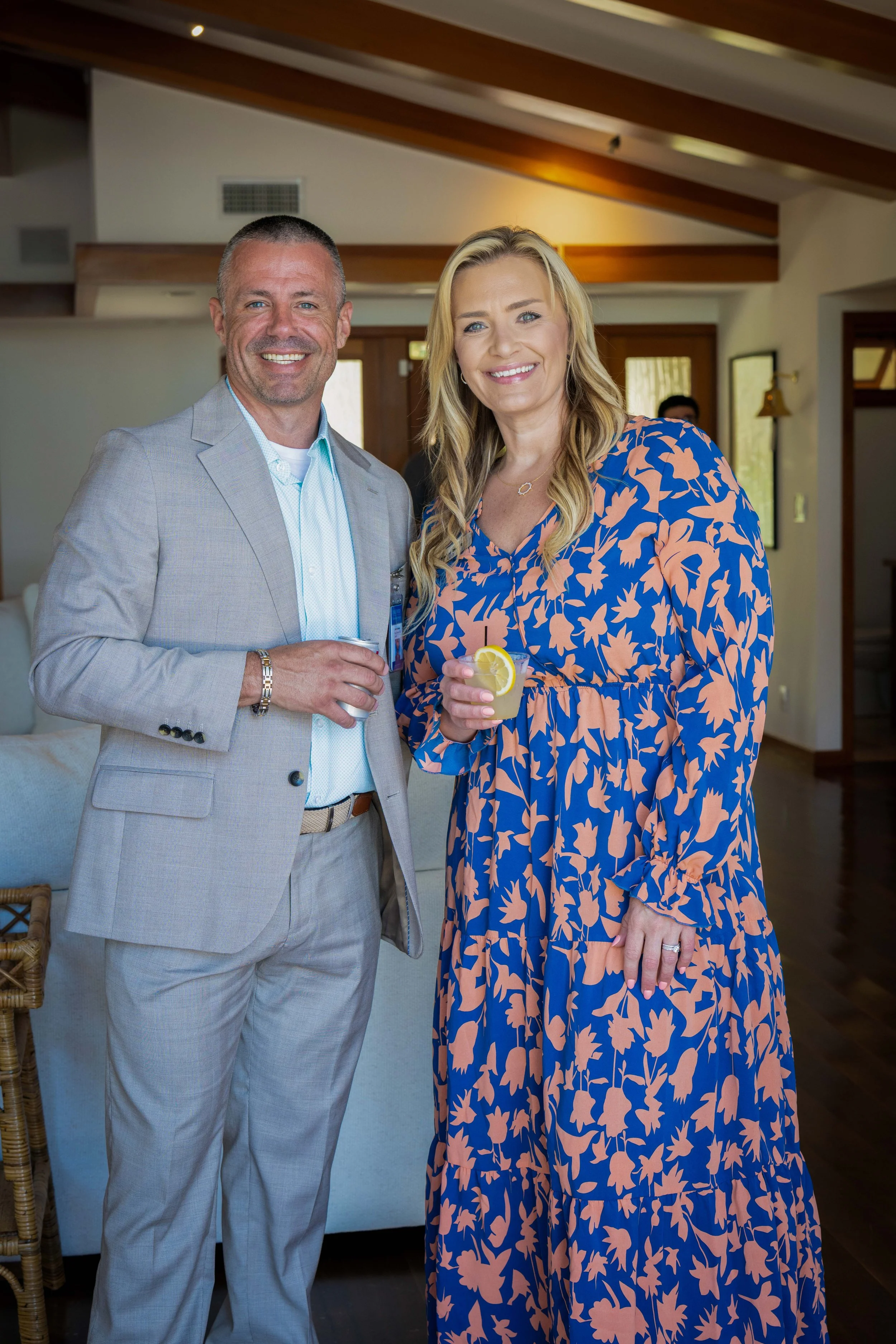 A man in a light gray suit and a woman in a blue and peach floral dress are smiling and holding drinks with lemon slices, standing inside a cozy, well-lit room with wooden ceiling beams and a dark floor.