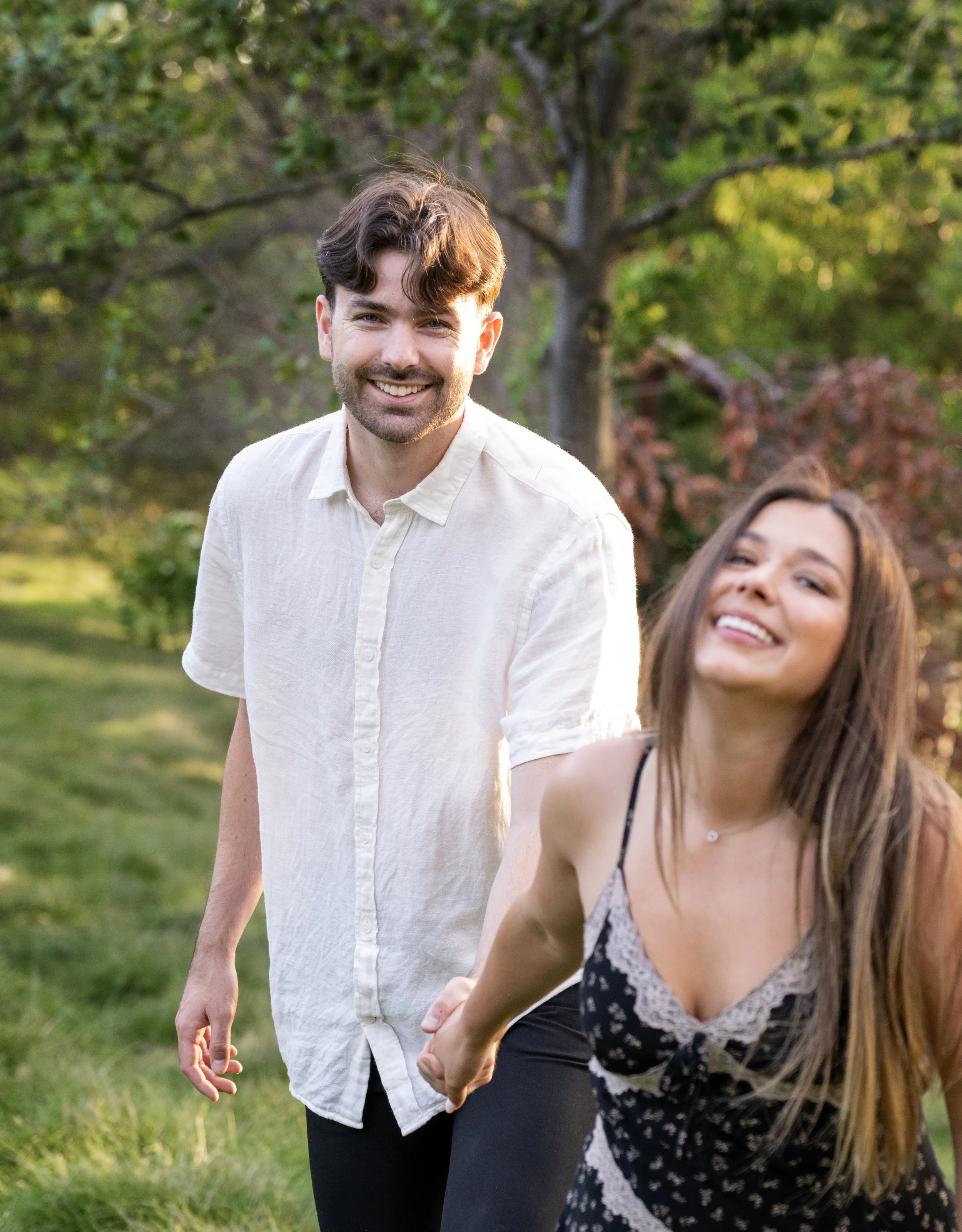 A smiling man with brown hair and a beard wearing a white shirt, holding hands with a woman with long brown hair and a patterned dress, outdoors in a park with green trees and grass.
