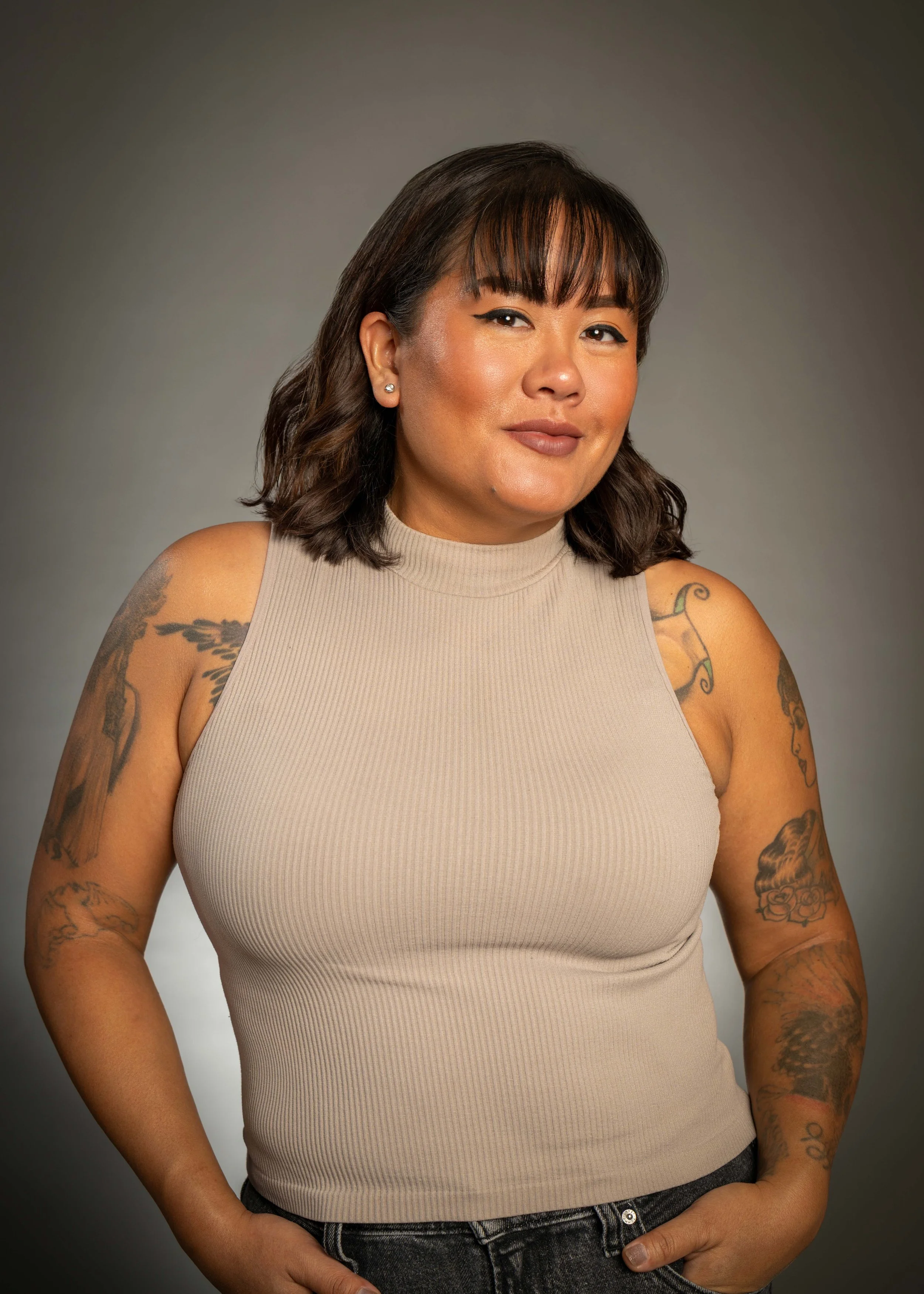 Portrait of a woman with shoulder-length dark wavy hair, wearing a beige sleeveless top, with tattoos on her arms, standing against a gray gradient background.