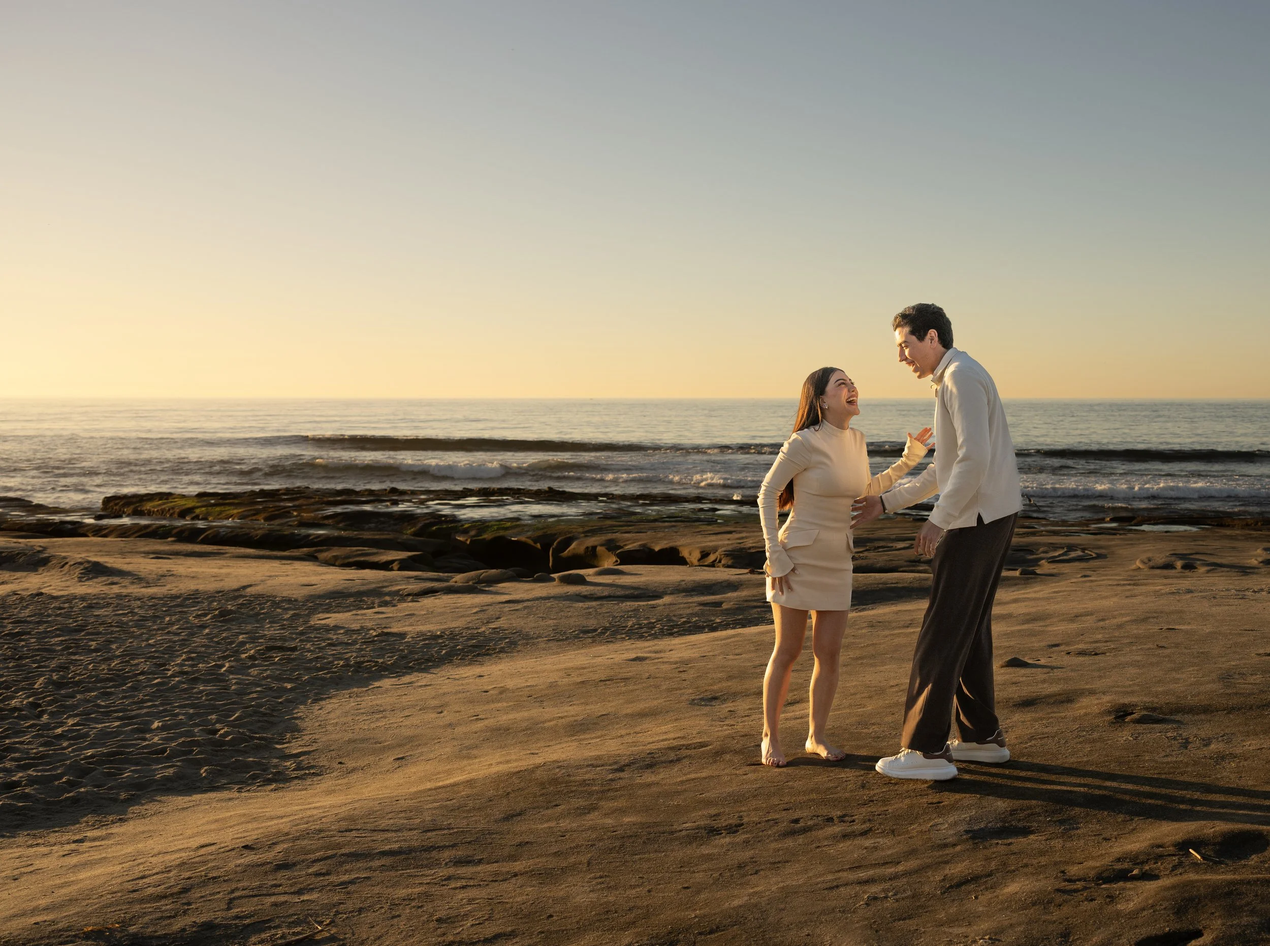 A man and woman smiling and holding hands while walking barefoot on a sandy beach during sunset.