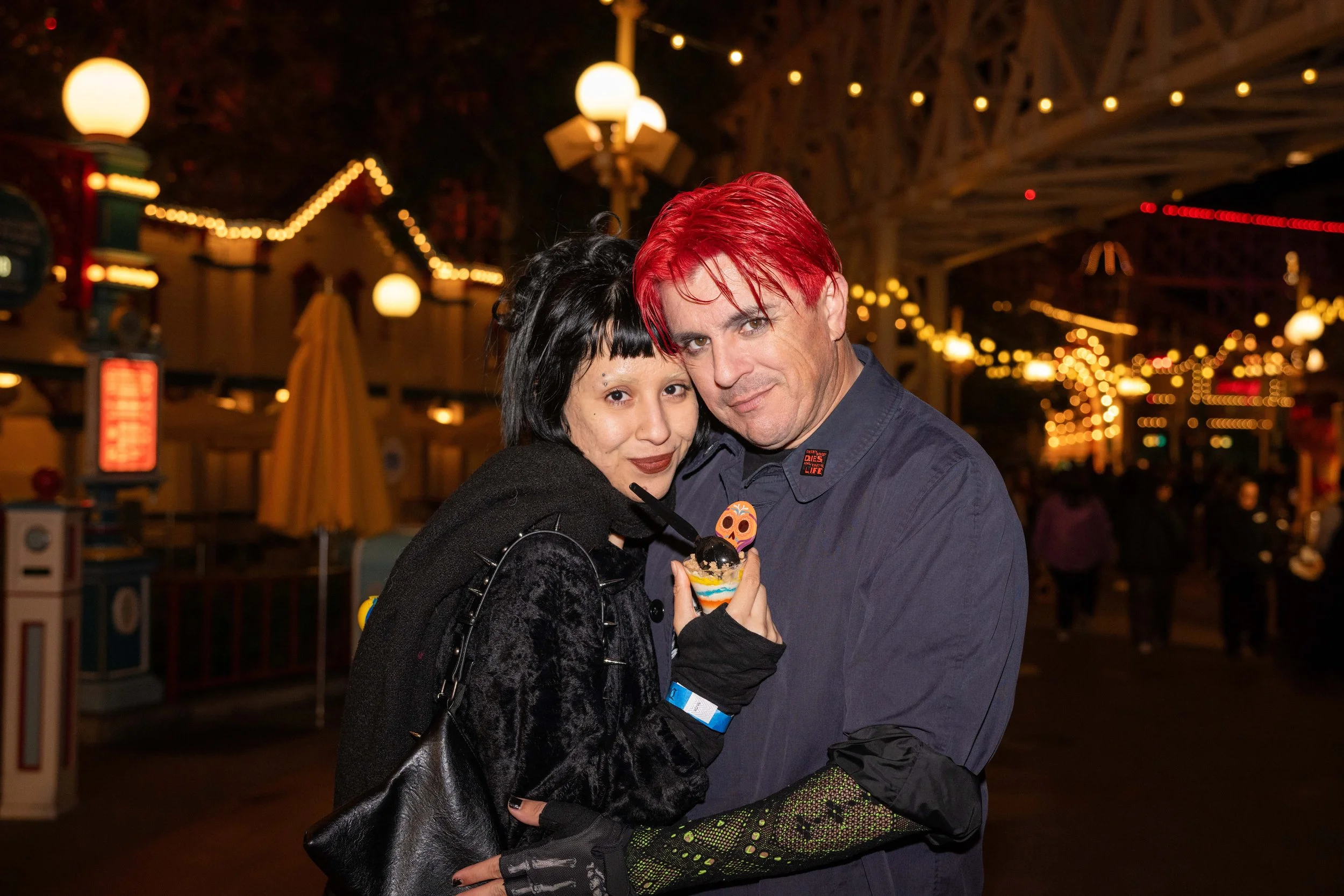 A woman and a man at an amusement park during the evening. The woman is holding a colorful ice cream and has black hair with a streak of white, wearing black gloves and a black coat, and the man has red hair, a blue shirt, and a button with a skull d