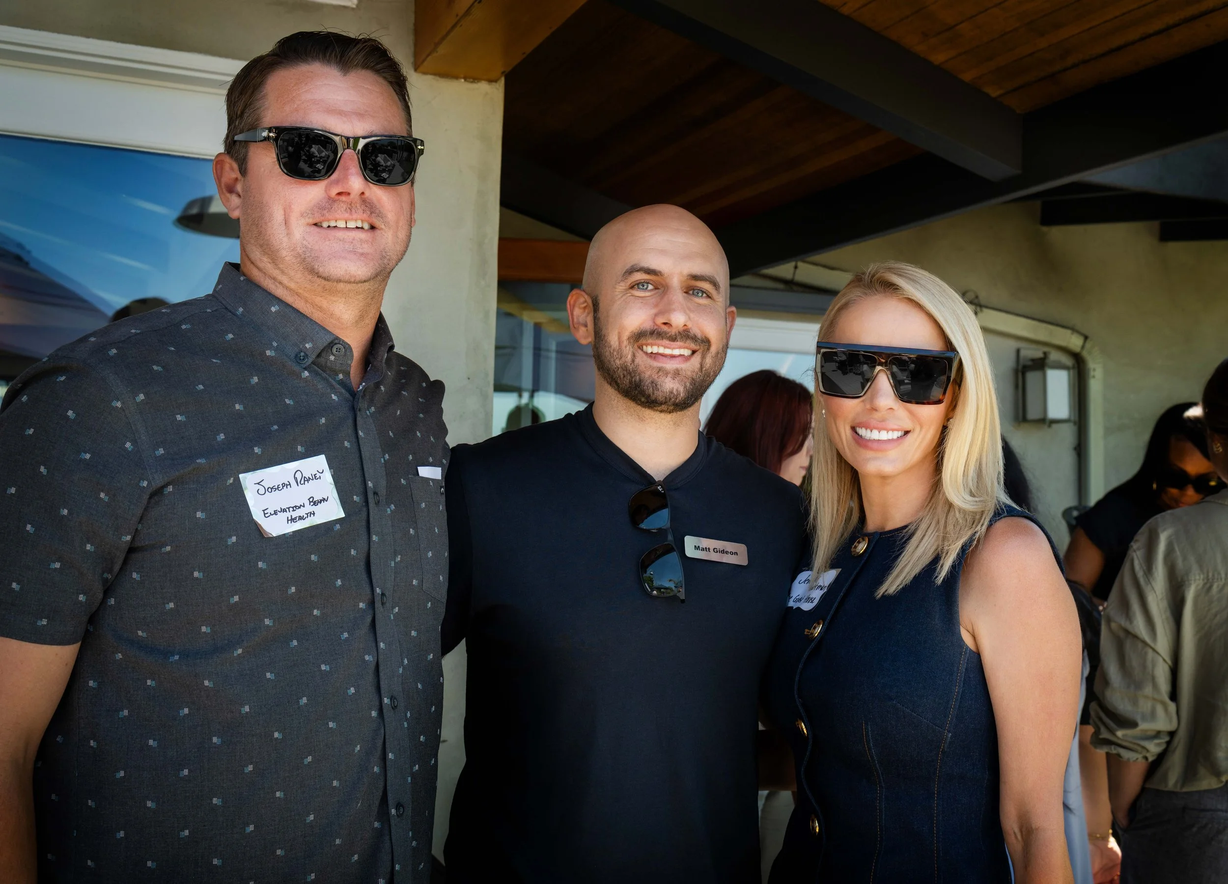 Three people attending an outdoor event, smiling at the camera. The man on the left is wearing sunglasses, a dark button-up shirt with small patterns, and a name tag. The man in the middle is bald, has a beard, and is wearing a black shirt, with sung