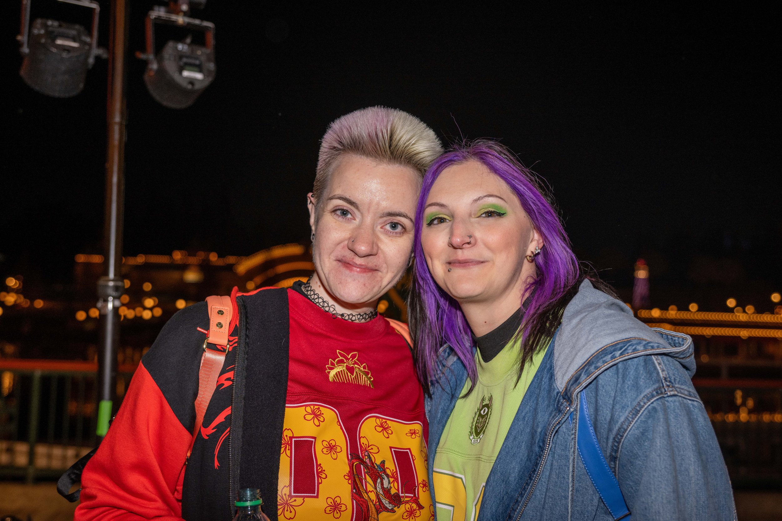 Two women with colorful hair and tattoos taking a selfie outdoors at night with city lights and a bridge in the background.