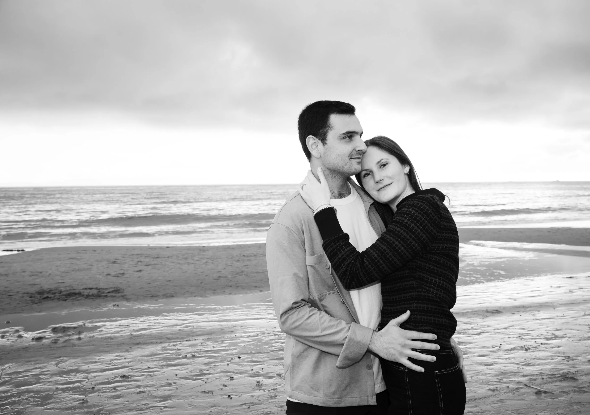 A black and white photo of a couple embracing on a beach with ocean waves in the background.