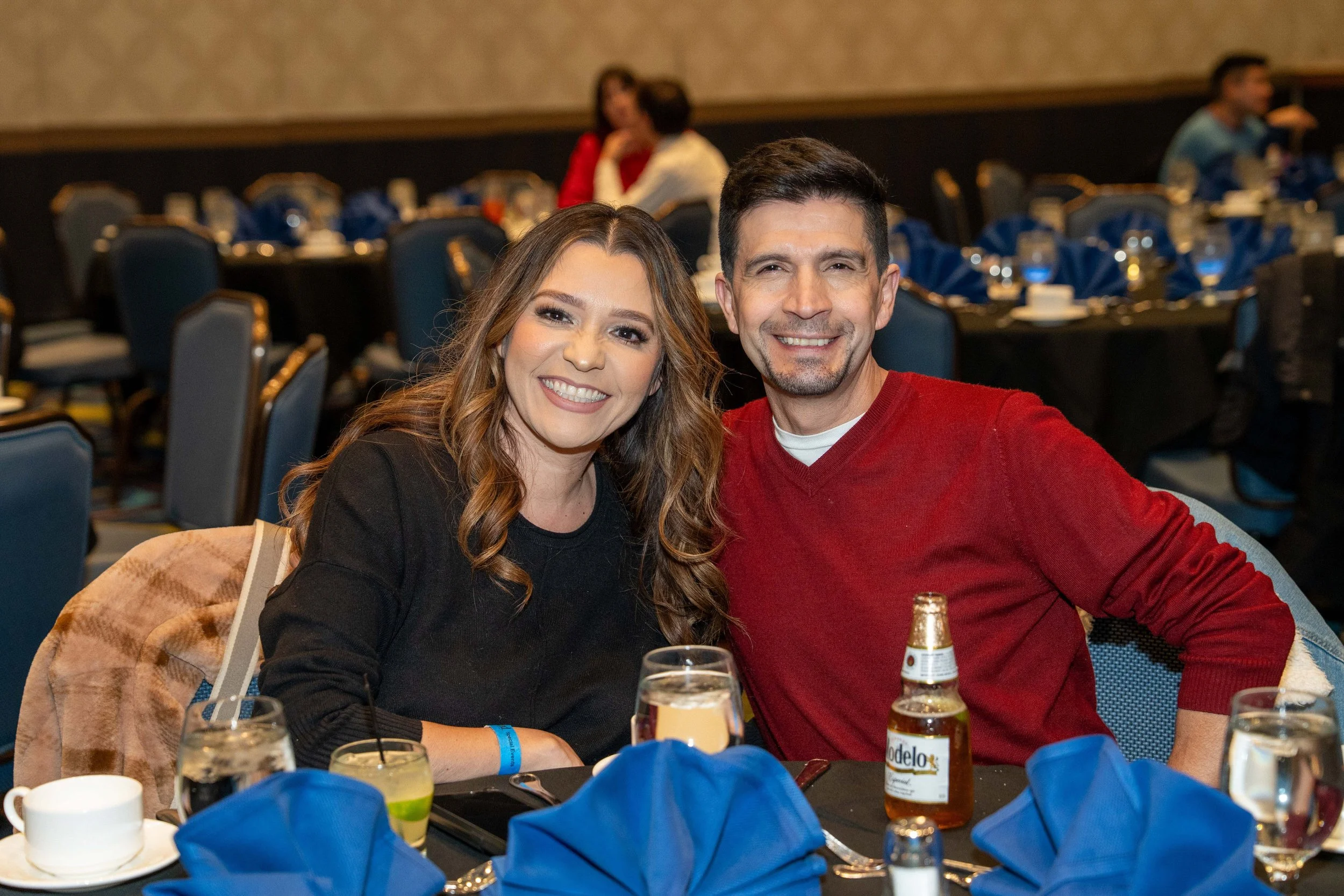 A smiling man and woman sitting together at a dining table during a social event, with plates, drinks, and glasses on the table.