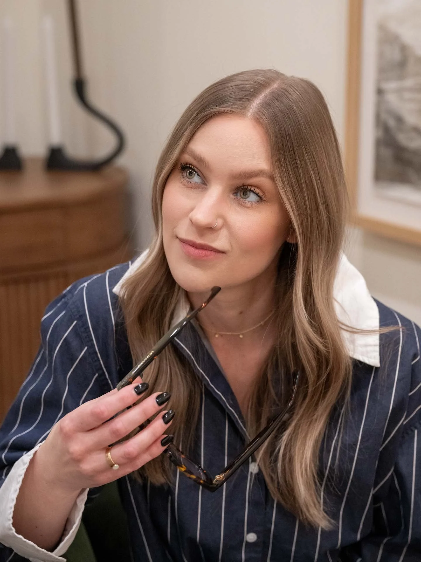 A woman with long, wavy hair, wearing a navy blue pinstripe shirt, holds a pair of glasses with her hand, which has black nail polish, and is looking to the side.