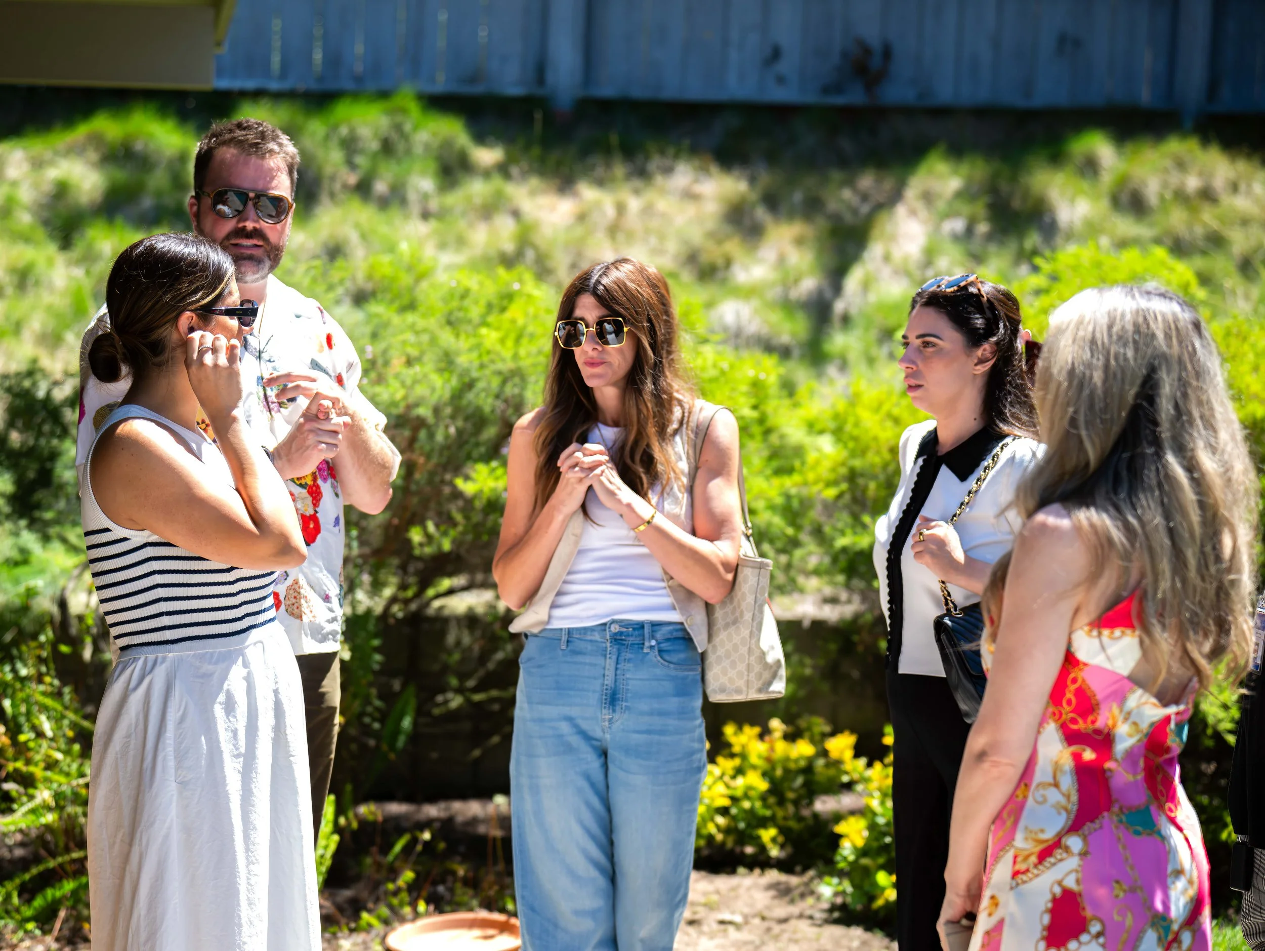 Group of five women and one man standing outside in a garden, engaged in conversation, some wearing sunglasses and casual summer clothing.