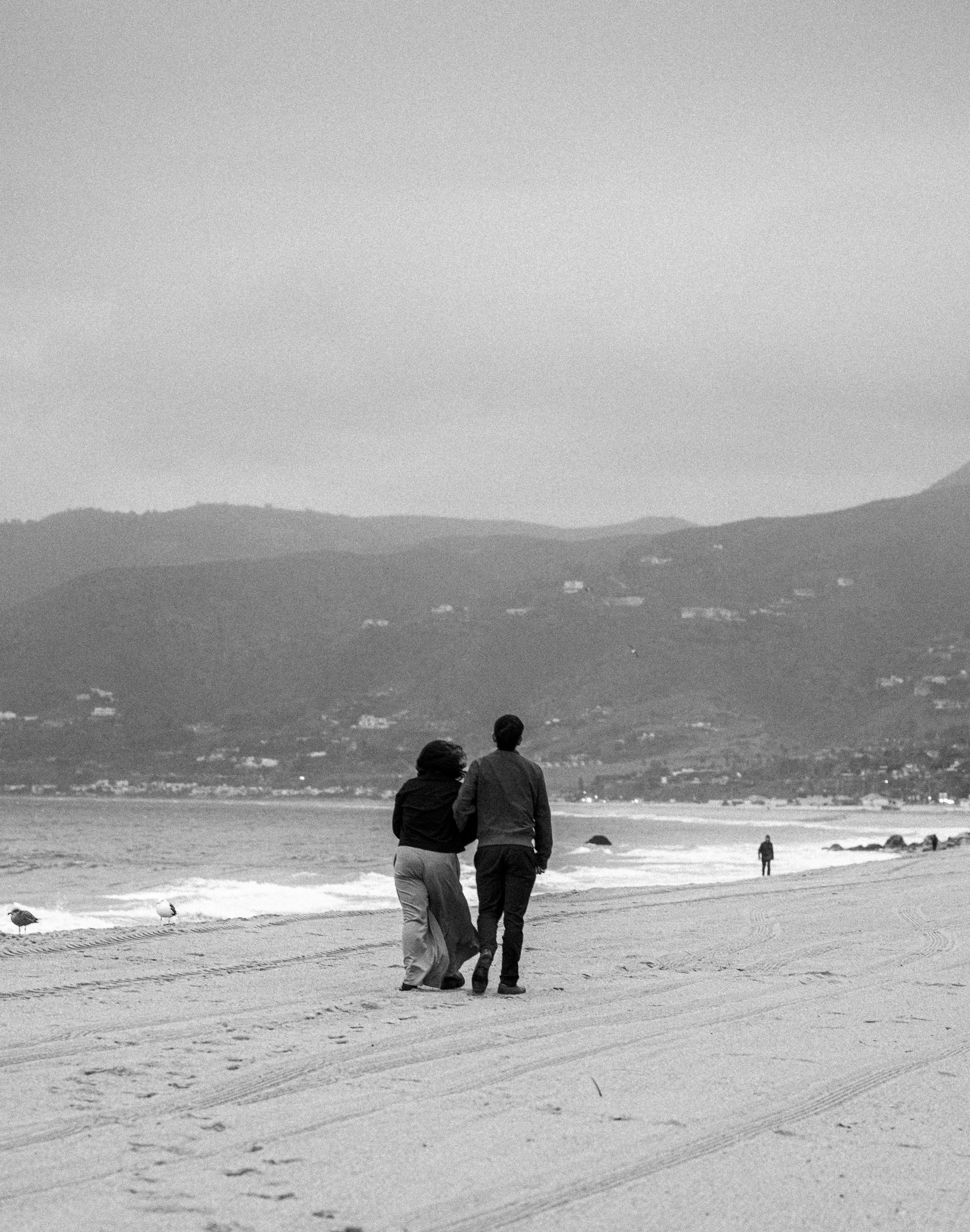 Two people walk along a sandy beach with their backs to the camera, with mountains visible in the background and a few other figures further along the shoreline.