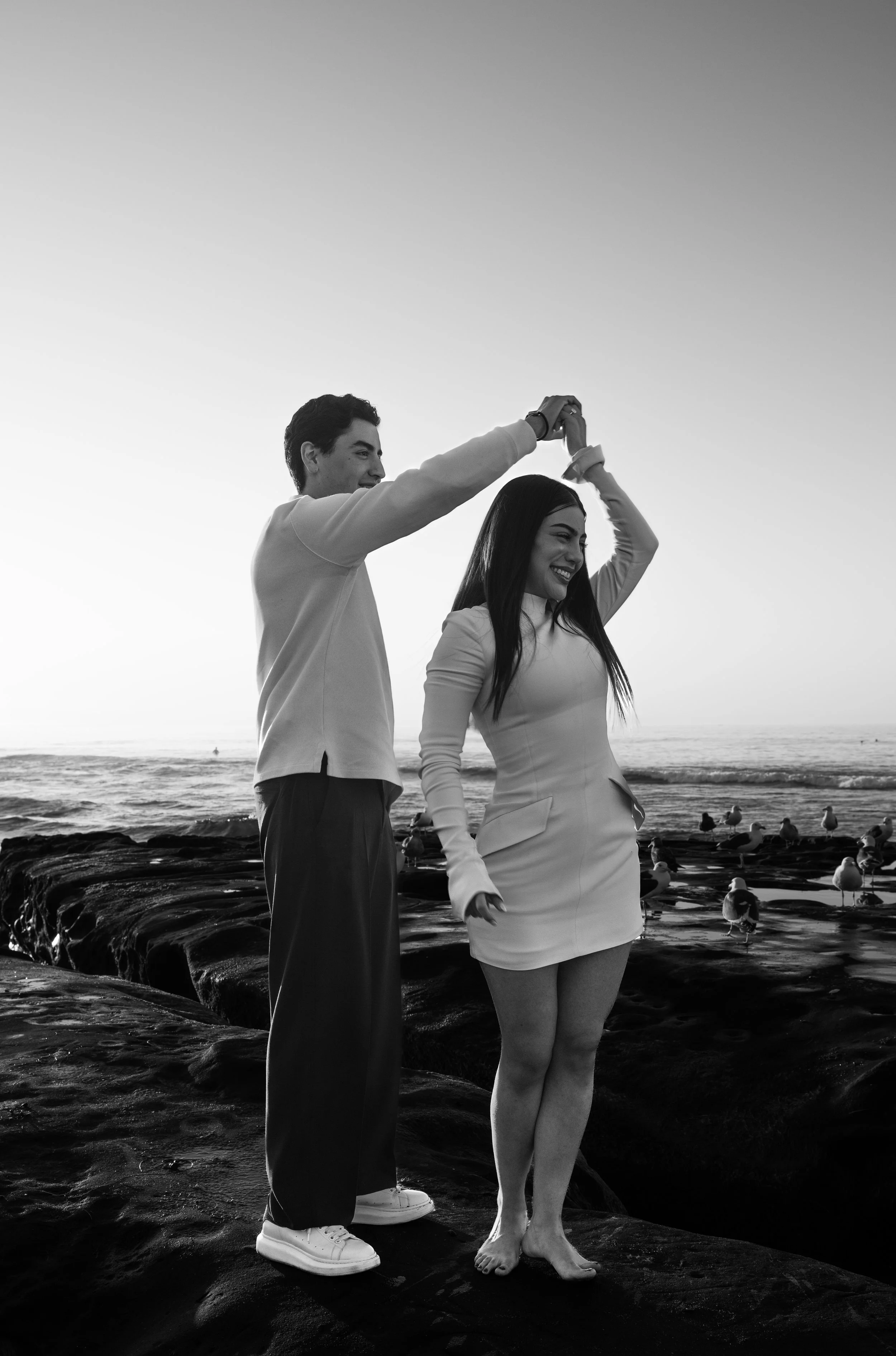 A young man and woman standing on rocks at the beach, holding hands above their heads in a dance pose, with seagulls and the ocean in the background, in black and white.
