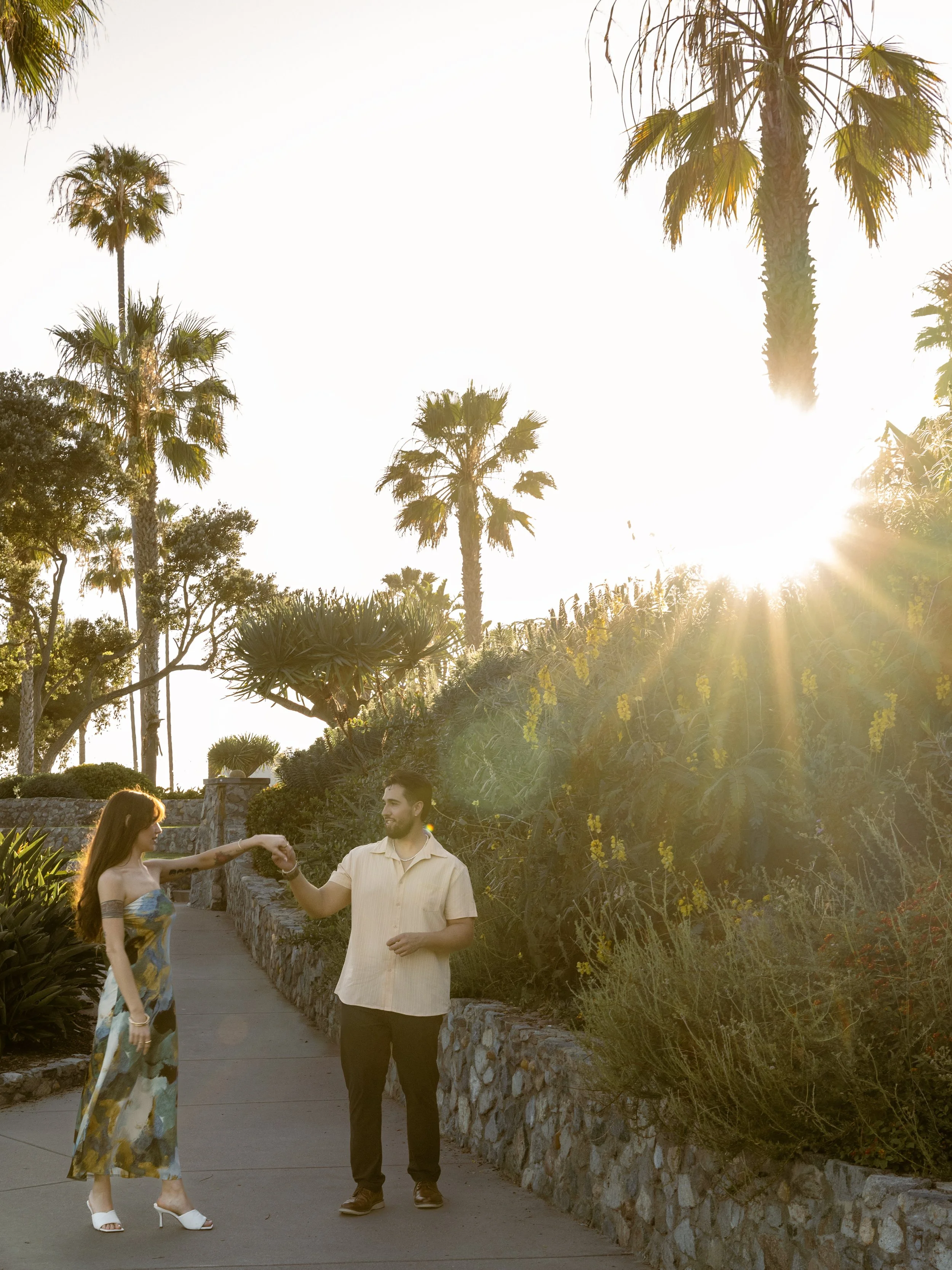 A woman and a man are holding hands and dancing on a sidewalk beside a stone wall with lush green plants and yellow flowers. Tall palm trees and the bright sun are visible in the background.