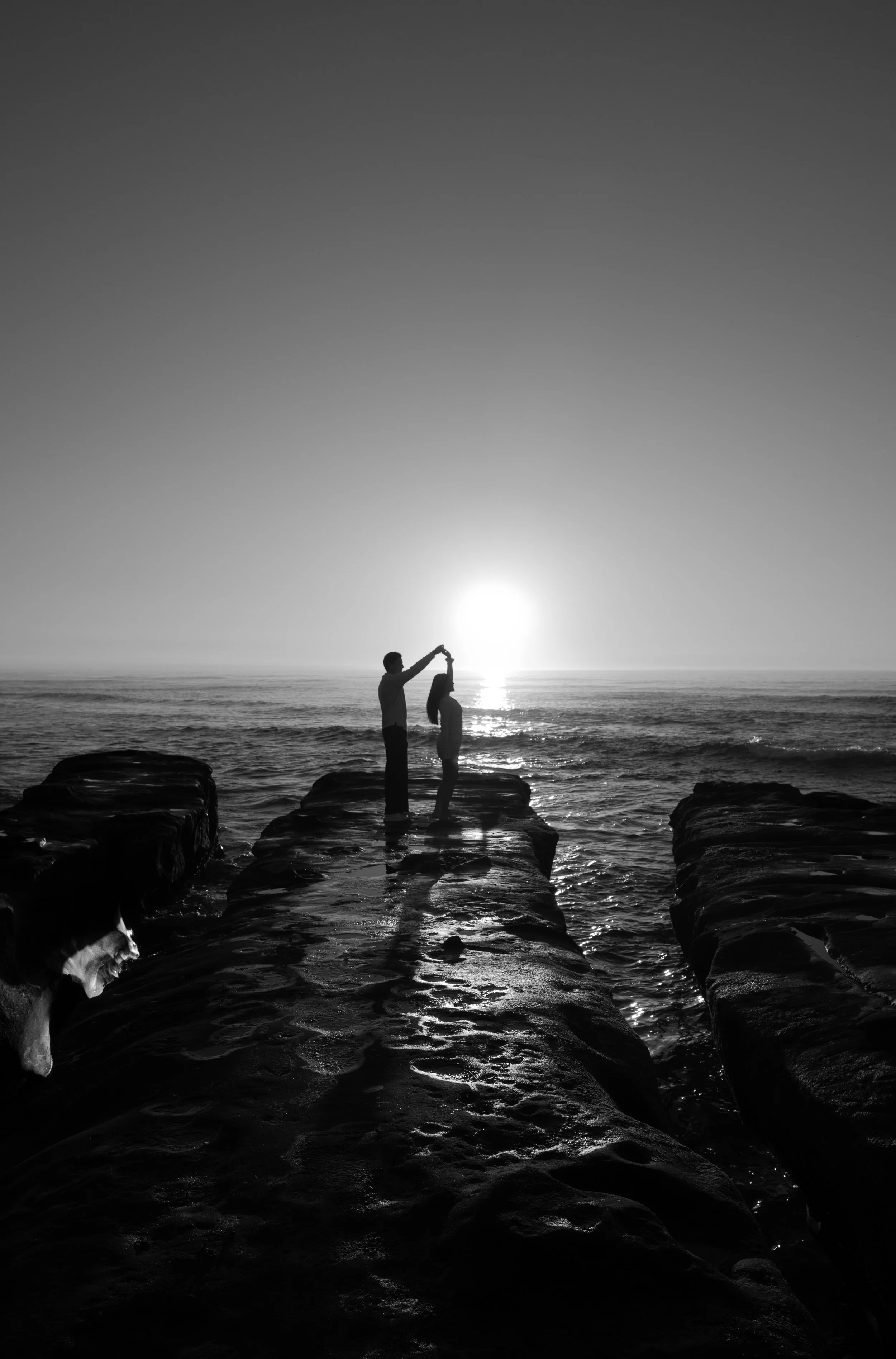 Silhouettes of two people on rocks at a lake during sunset, with one person taking a photo of the other, and the sun near the horizon.