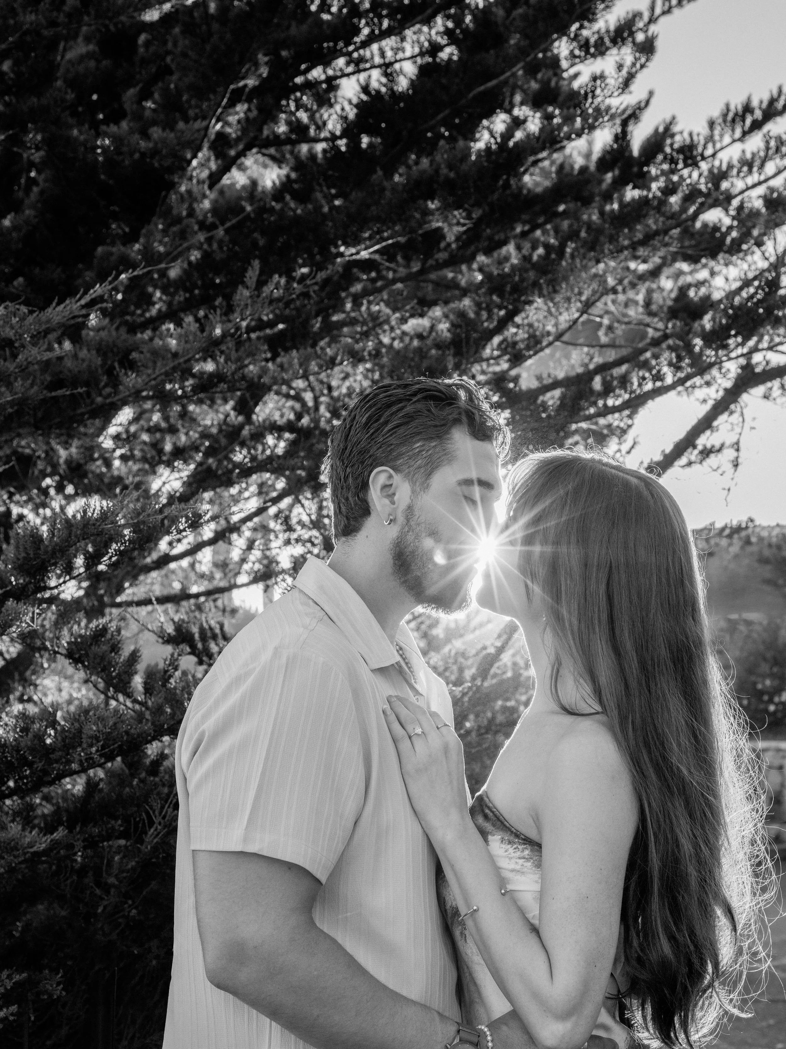 A black and white photo of a couple performing a romantic kiss outdoors with trees in the background and sunlight creating a starburst effect.