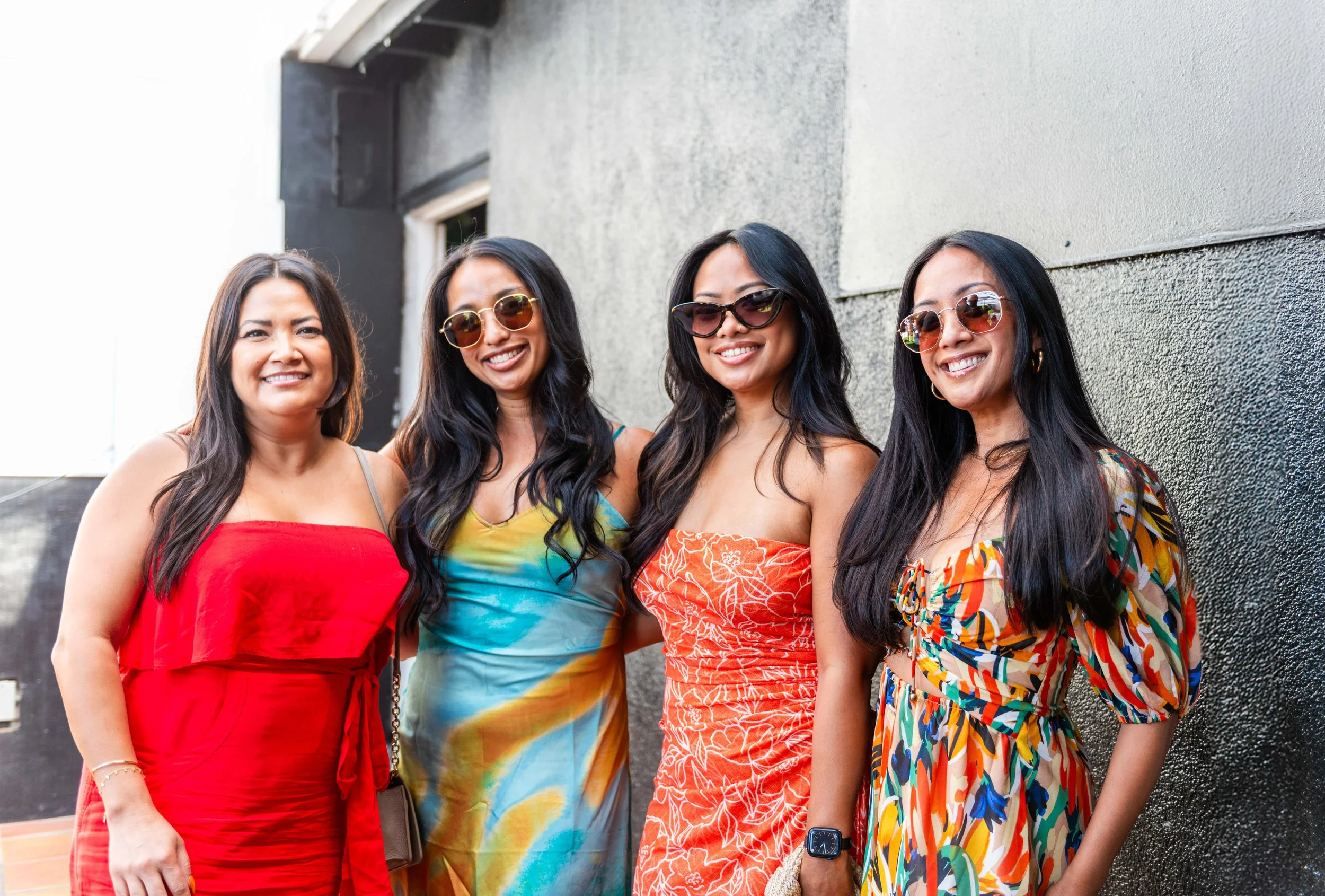 A group of four women standing close together outdoors against a textured wall, smiling, wearing colorful summer dresses and sunglasses.