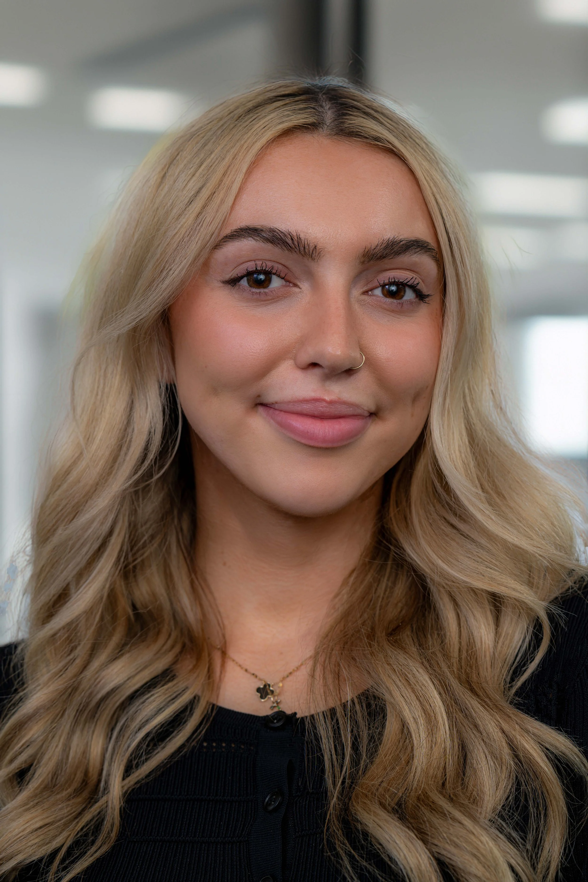 Close-up portrait of a young woman with long, wavy blonde hair, wearing a black top and a delicate necklace, standing indoors with blurred ceiling lights in the background.