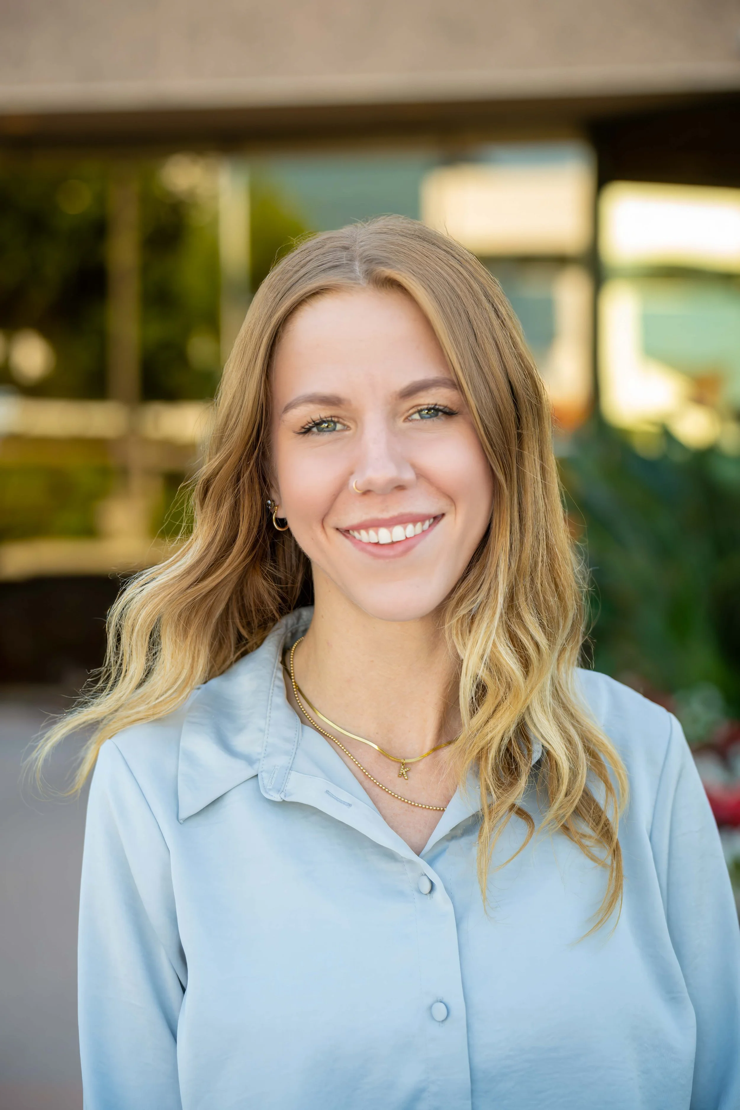 A young woman with long wavy blonde hair smiling outdoors in front of a blurred background of trees and a building.