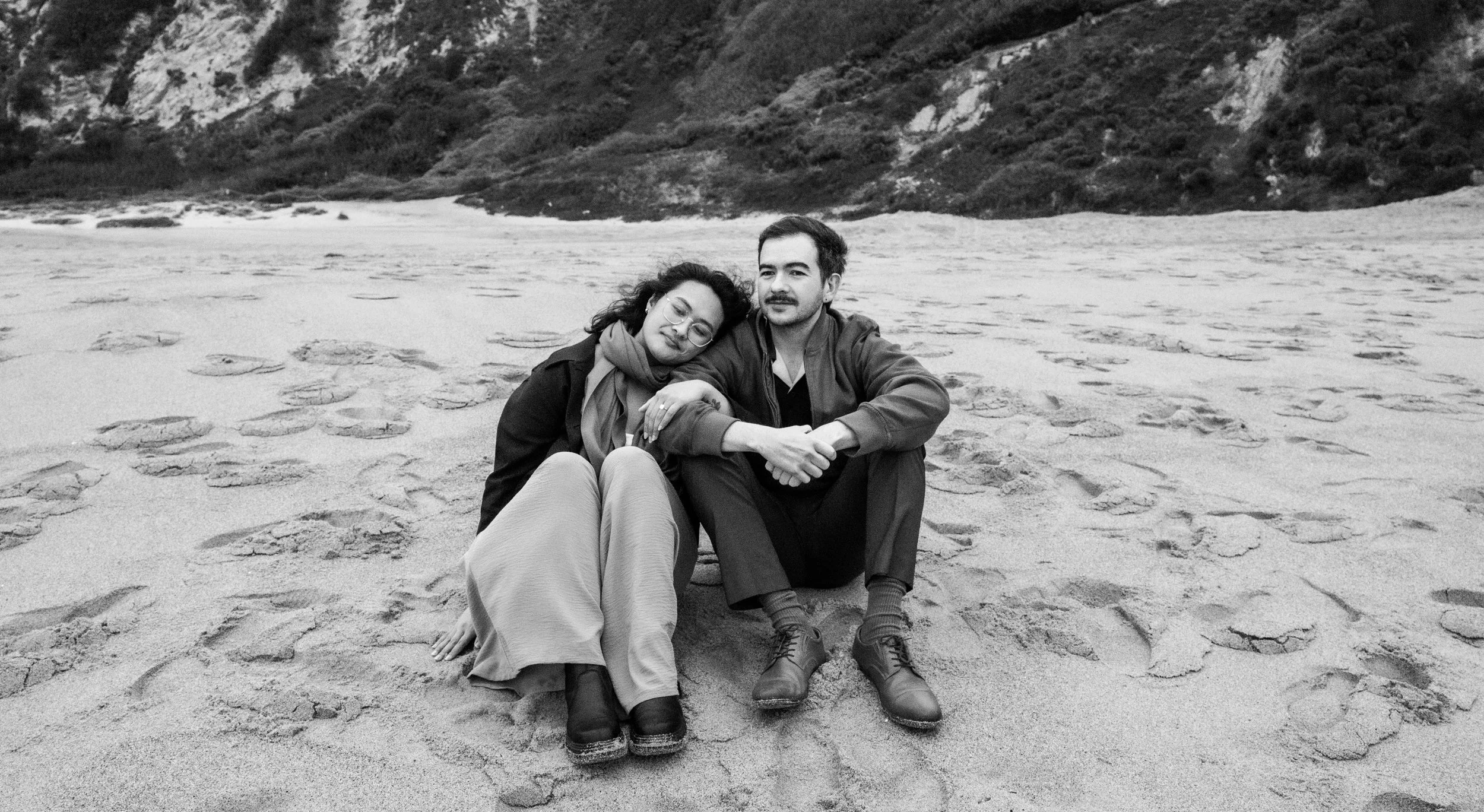 A black and white photo of a couple sitting on a sandy beach, with one woman resting her head on the man's shoulder. They are surrounded by footprints, with cliffs and vegetation in the background.