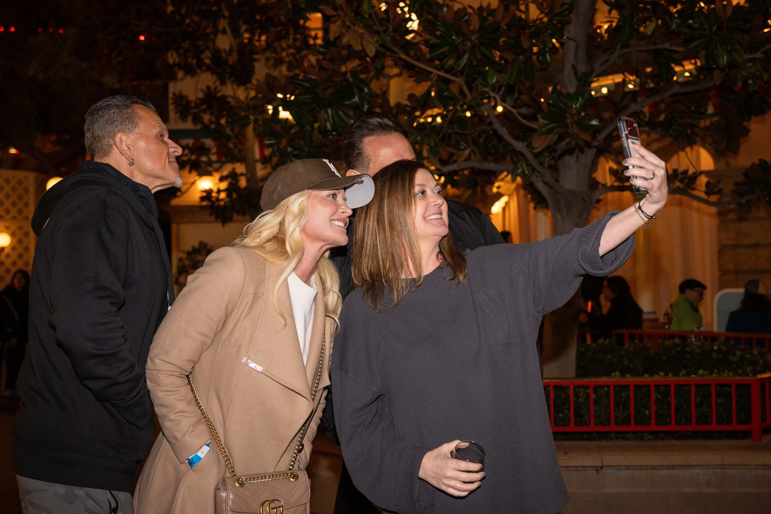 Four people taking a selfie outdoors at night. The group includes two women and two men smiling, with a background of trees and warm yellow lighting.