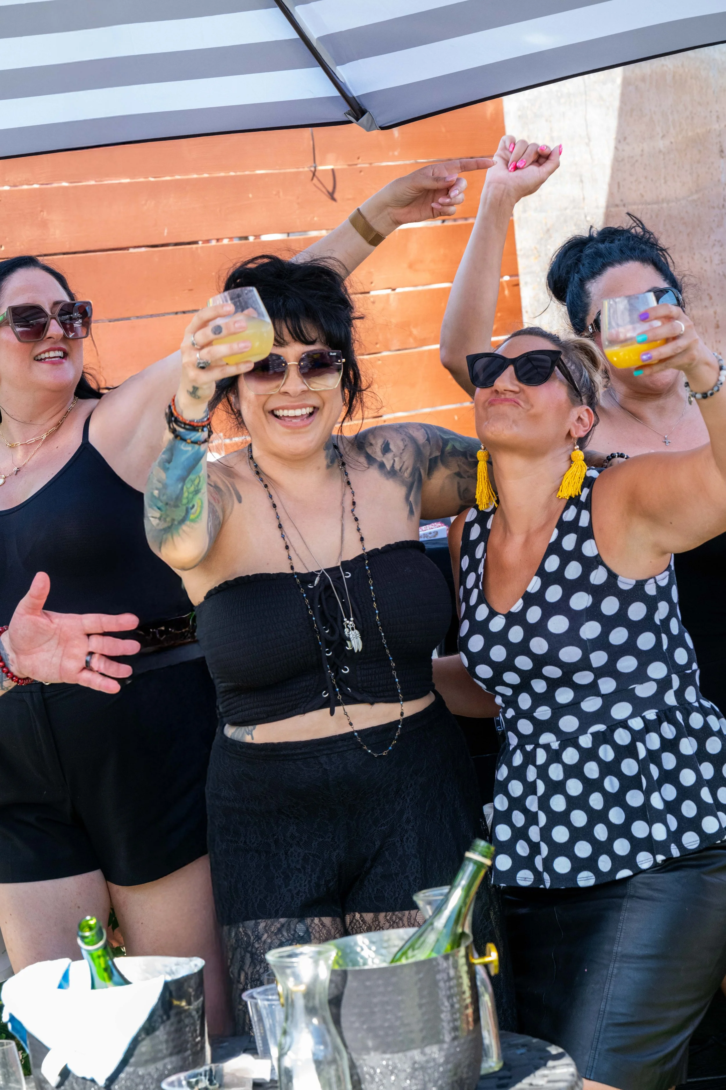 A group of five women celebrating outdoors, holding drinks, wearing sunglasses, and smiling. They are gathered around a table with bottles and a ice bucket, with a wooden fence in the background.
