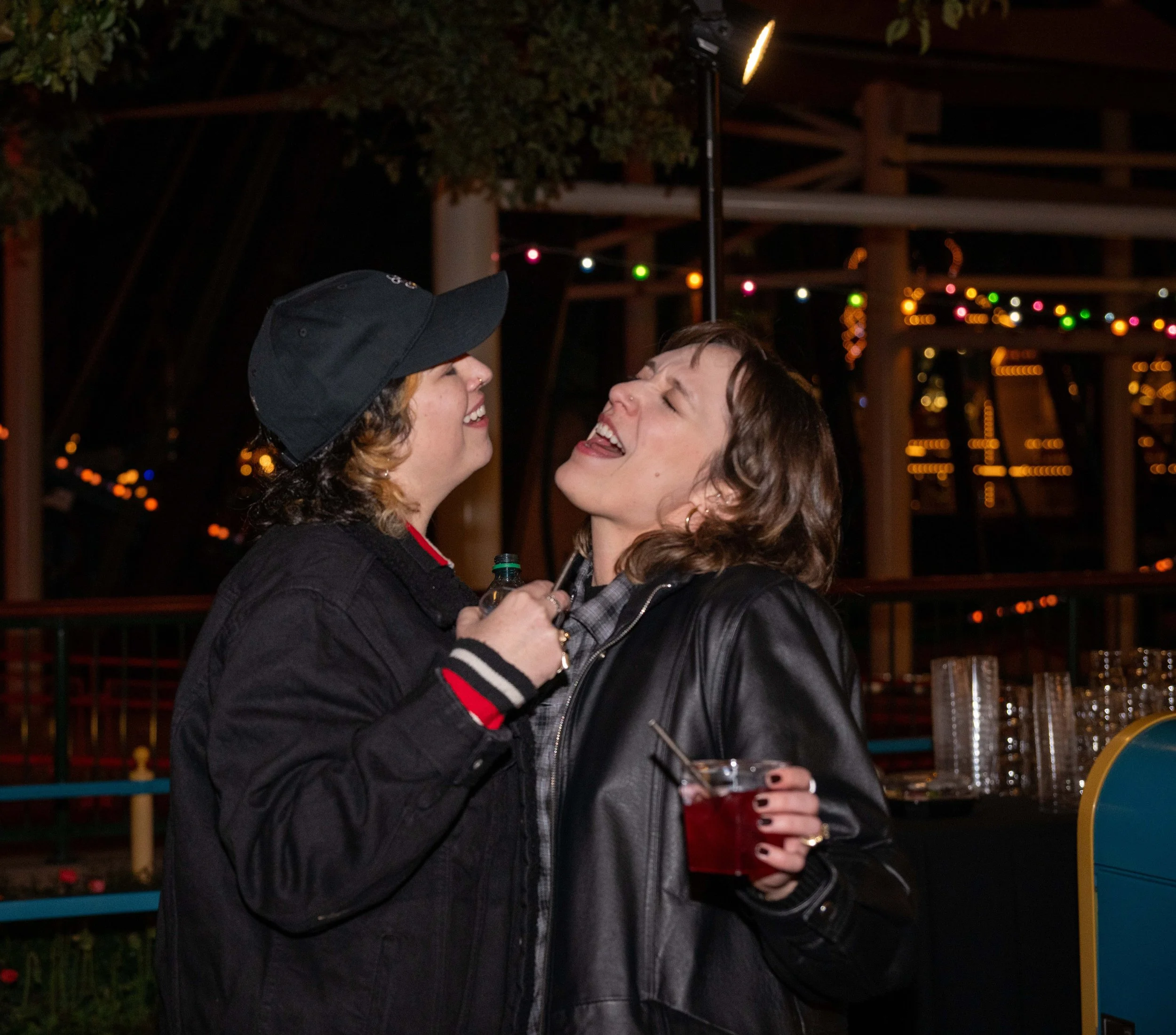 Two women smiling and laughing inside a festive setting with colorful string lights and wooden structures in the background. One woman is wearing a black hat and the other has curly hair, holding a drink.