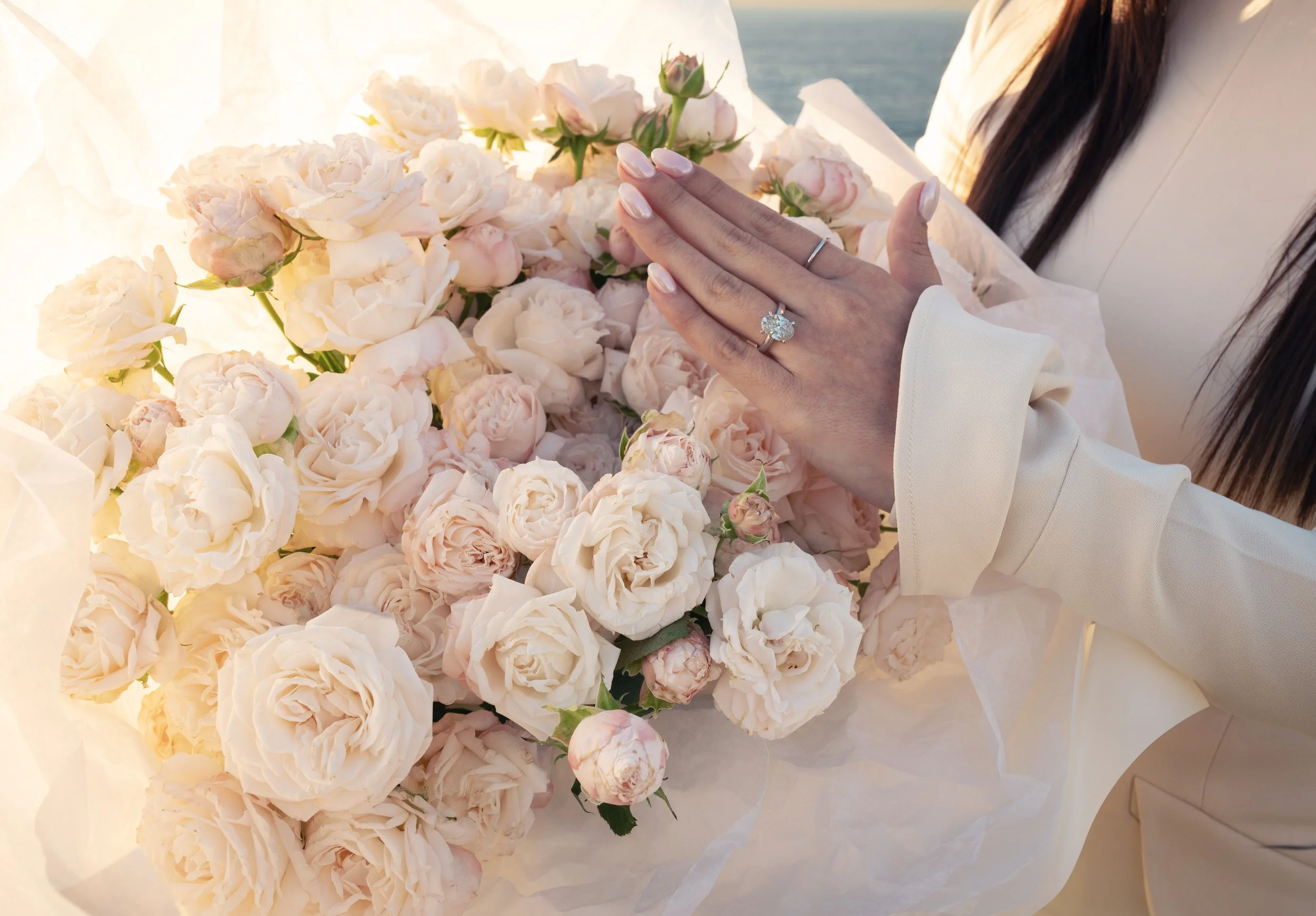 A person with dark hair, wearing a white outfit, is holding a large bouquet of pale pink and cream peonies. The person is showing off a large, sparkling gemstone ring on their left hand. The background suggests a seaside or ocean view.