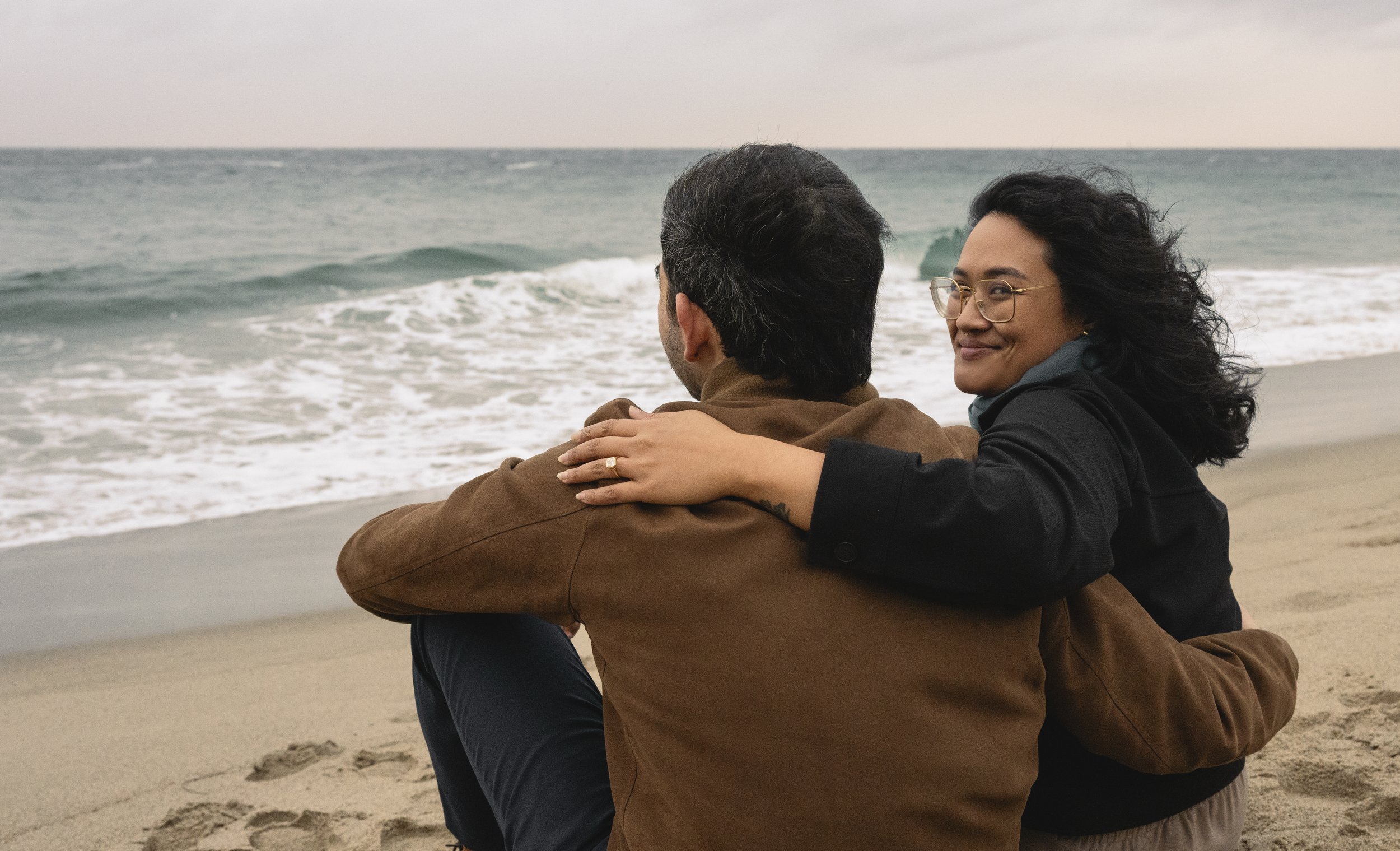 A woman and a man sit on the beach facing the ocean, with the woman smiling and resting her arm on the man's shoulder.