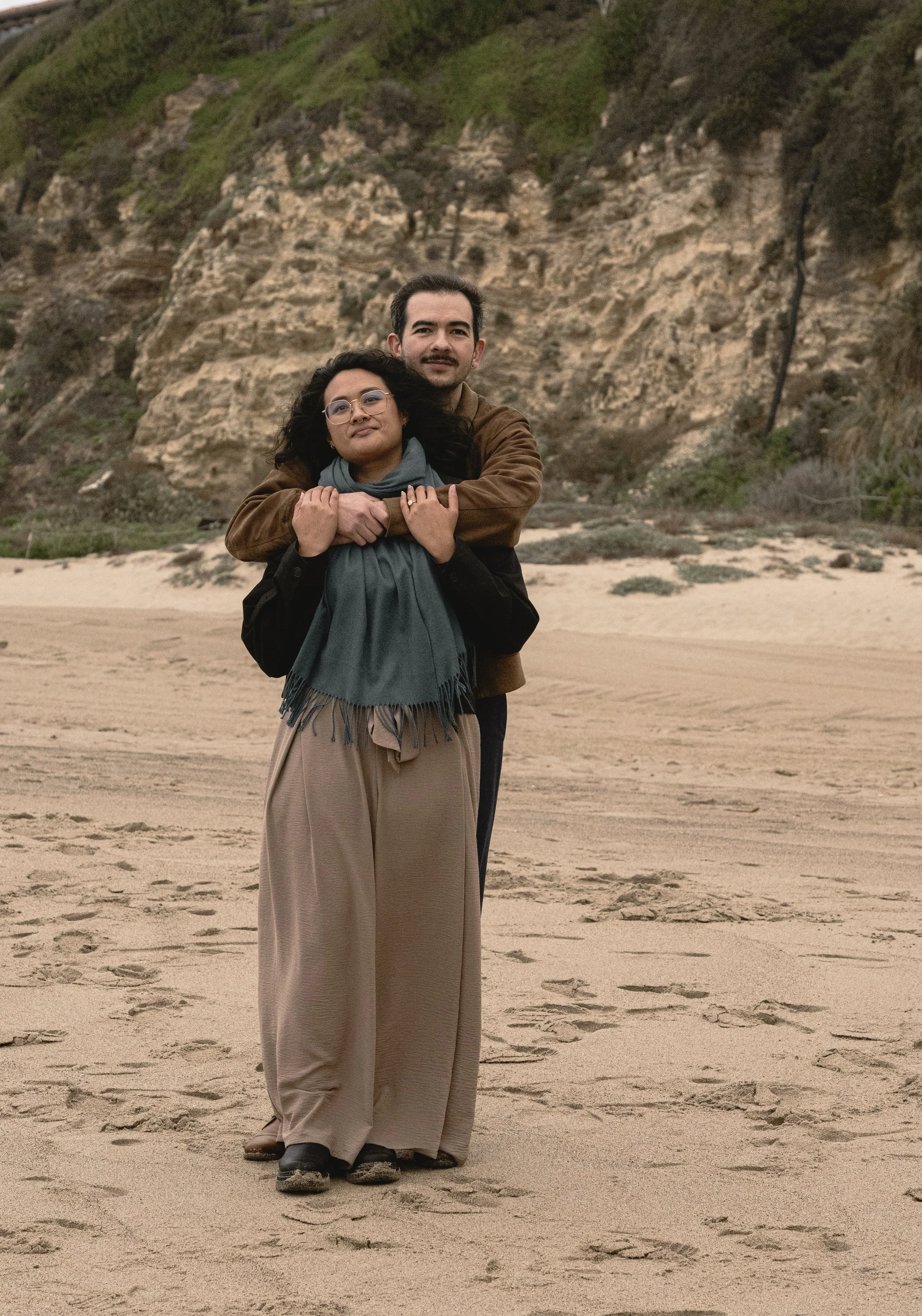 A man is giving a woman a piggyback ride on a sandy beach with cliffs and greenery in the background. The woman has curly dark hair and glasses, and the man has short dark hair and a mustache.