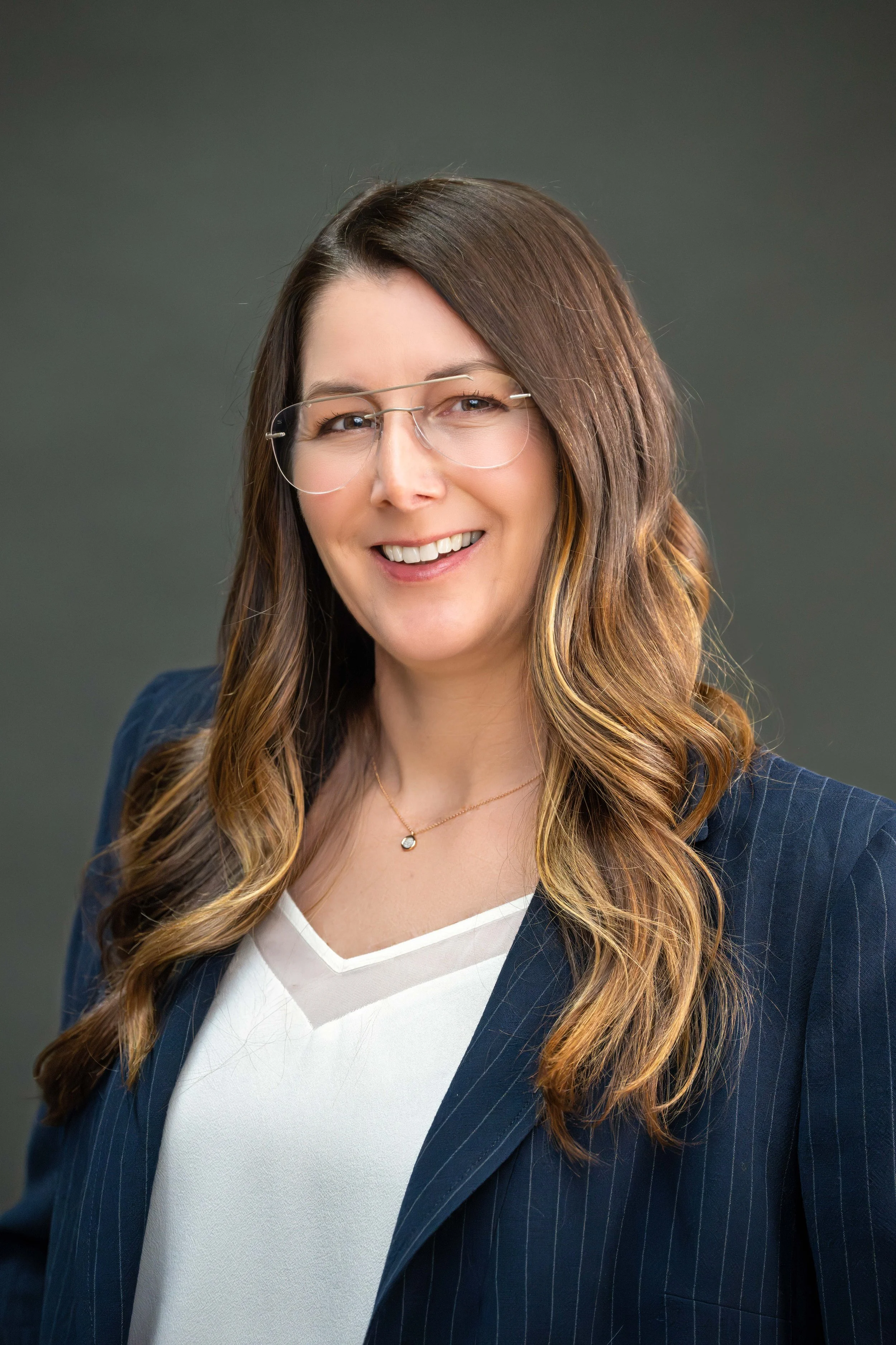 A smiling woman with long, wavy brown hair, wearing glasses, a white V-neck top, and a pinstripe navy blazer, standing against a gray background.