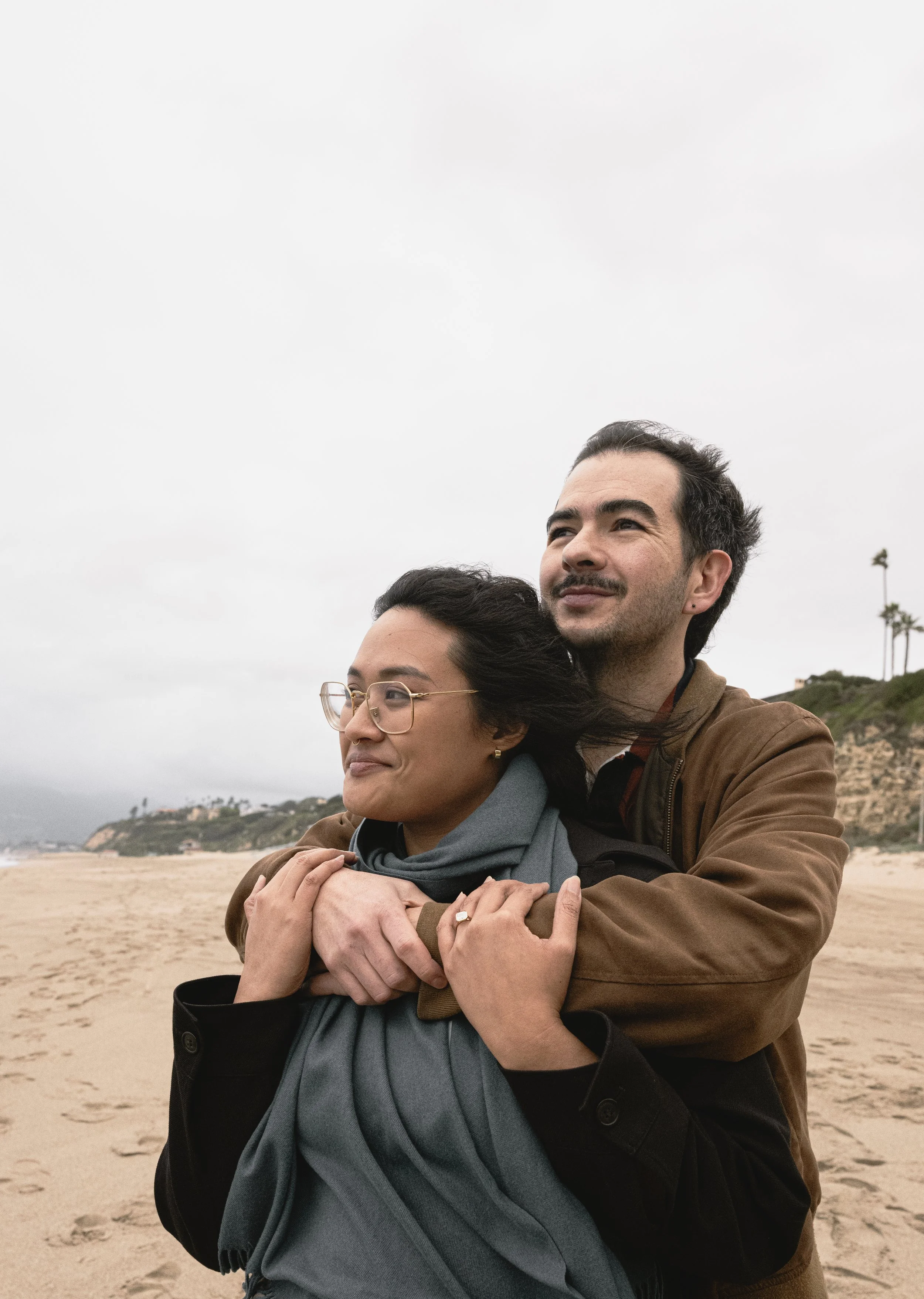 A man and woman embrace on a beach, with hills and palm trees in the background under a cloudy sky.