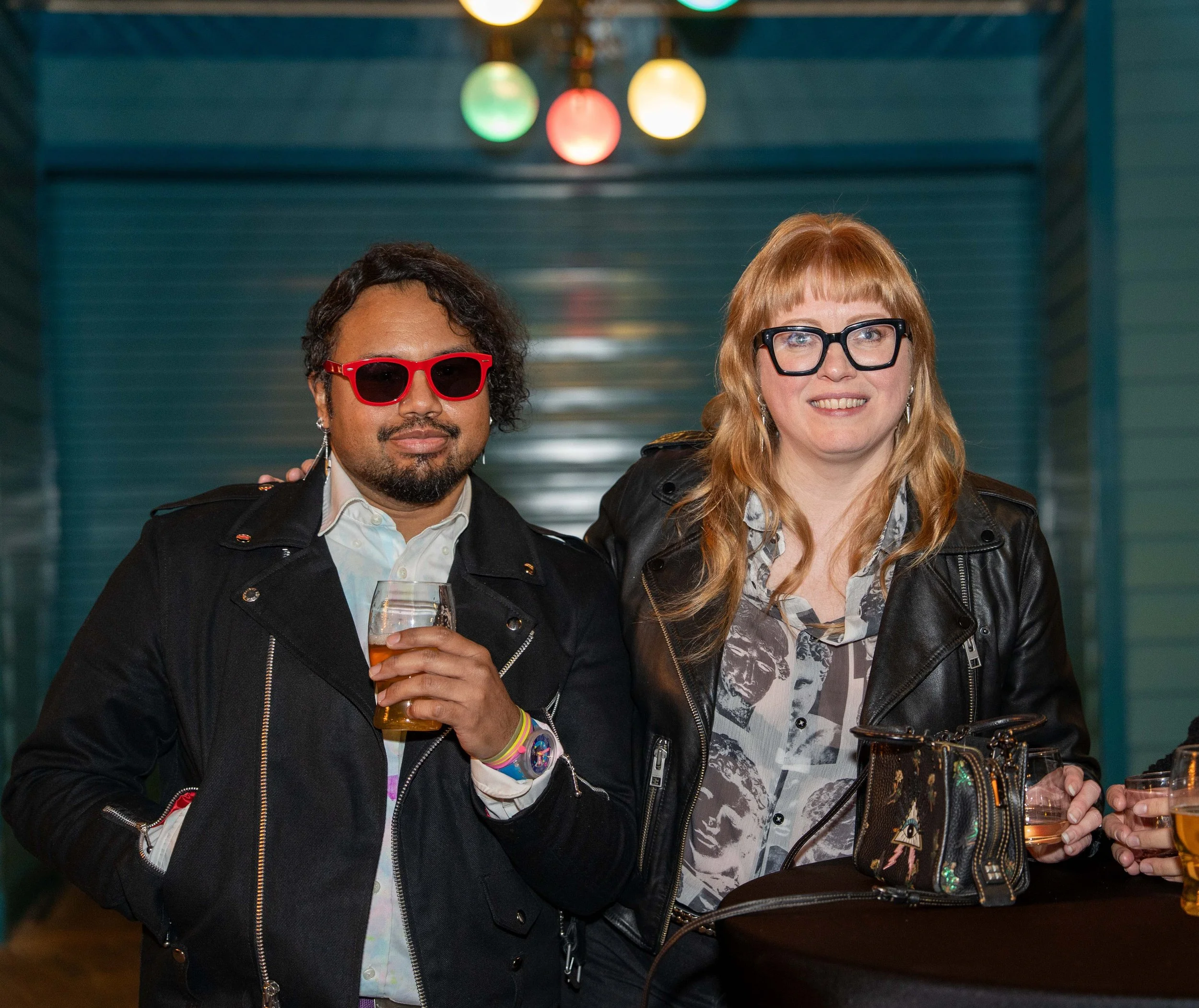 Two people at a social event holding drinks, one wearing red sunglasses and the other wearing black glasses, both smiling, with colorful lights in the background.